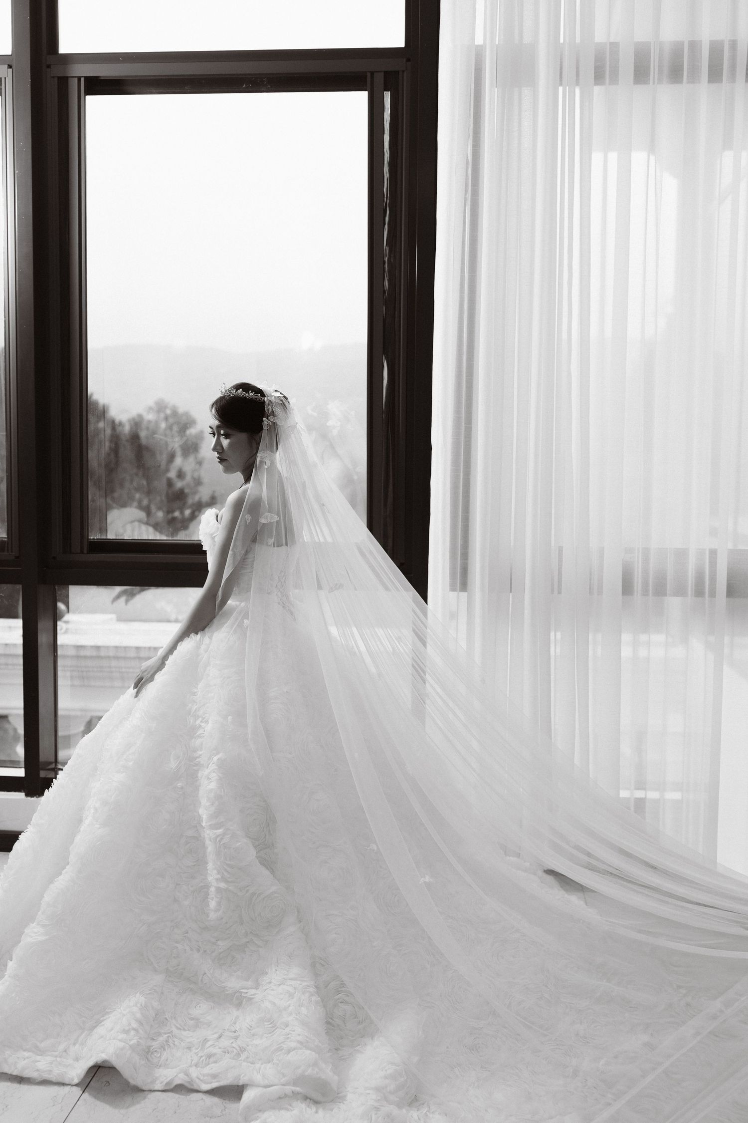 Black and white photo of a bride in a flowing lace gown and long veil standing by a large window.