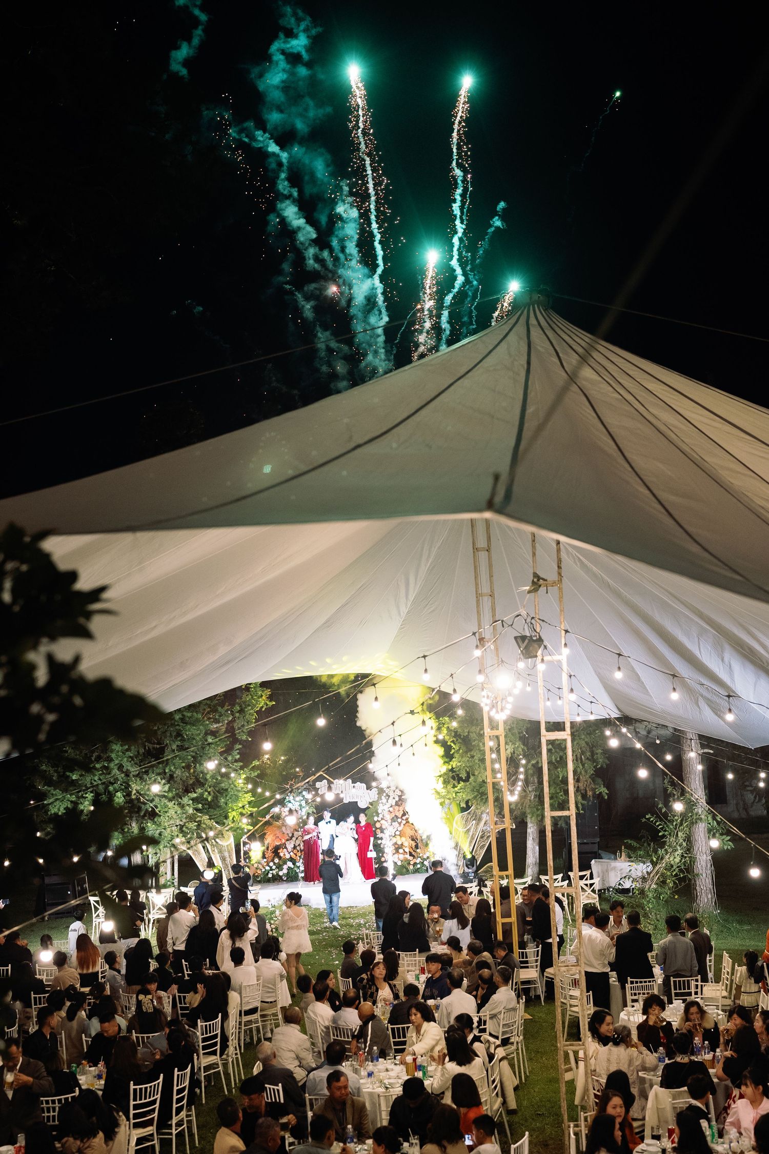 Outdoor wedding reception under a white tent at night with fireworks lighting up the sky above guests seated at tables.