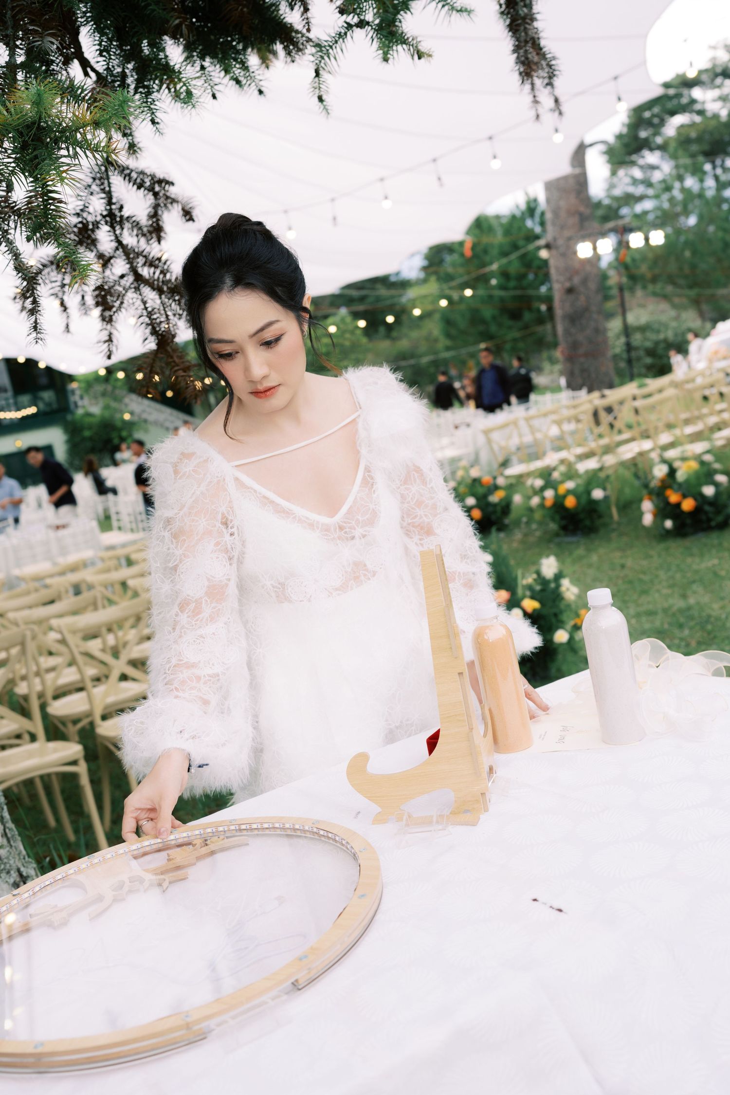 A bride in a white lace dress arranges items on a table at an outdoor wedding venue with string lights.