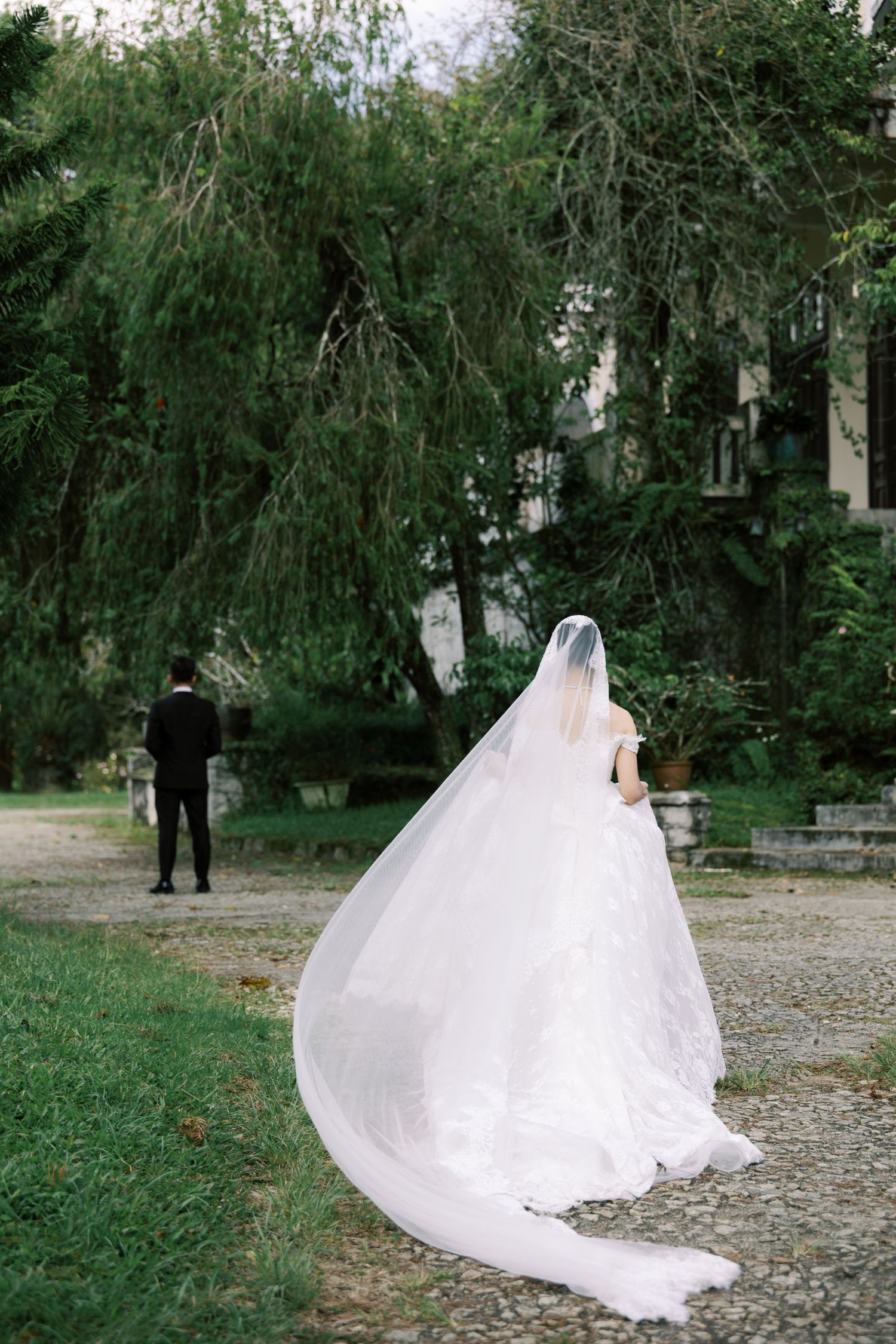 Bride in flowing white gown and veil walks toward groom in black suit in a lush green garden setting.