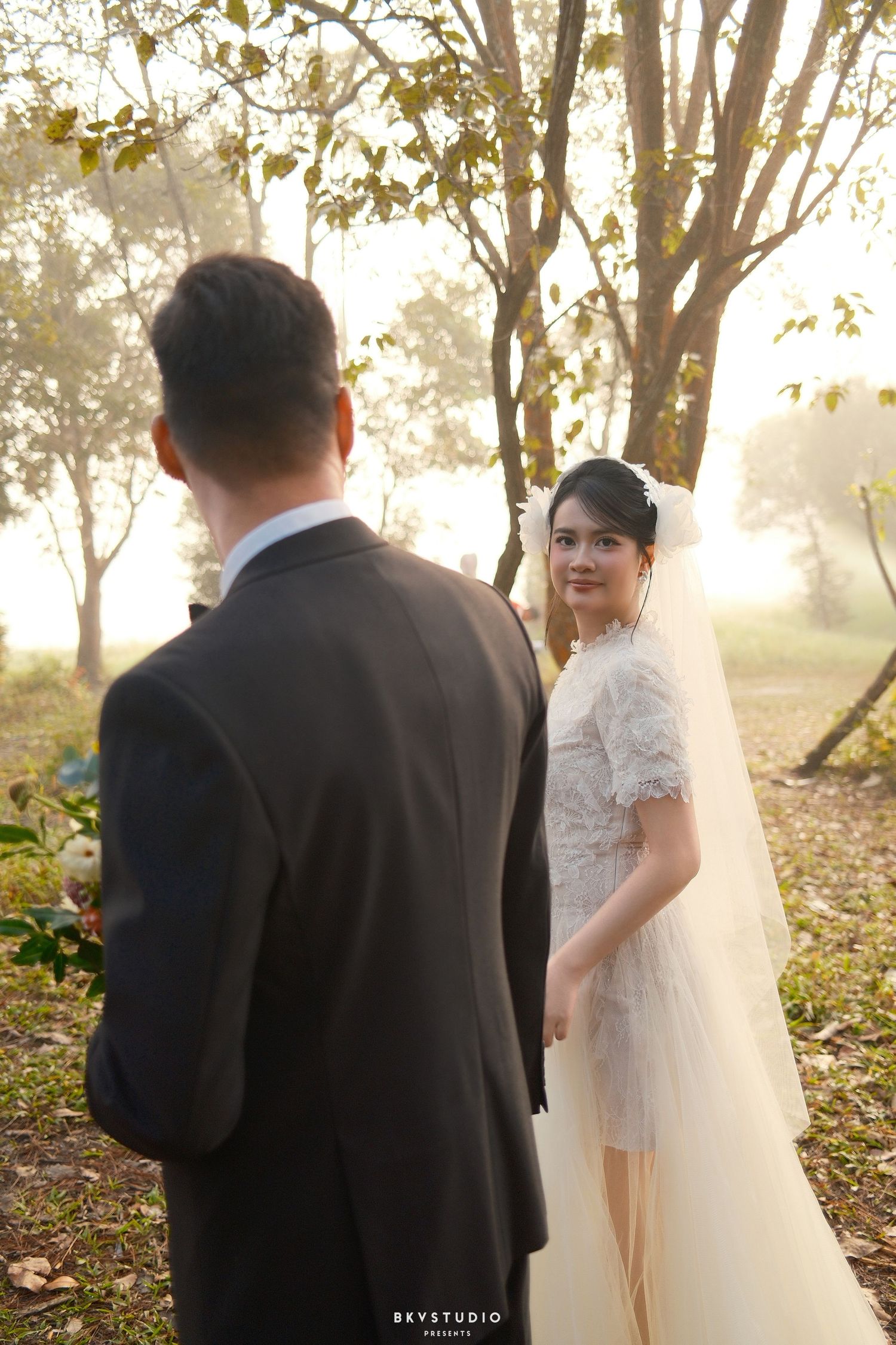 Bride in white lace dress glances back at groom among sunlit autumn trees during wedding photoshoot.