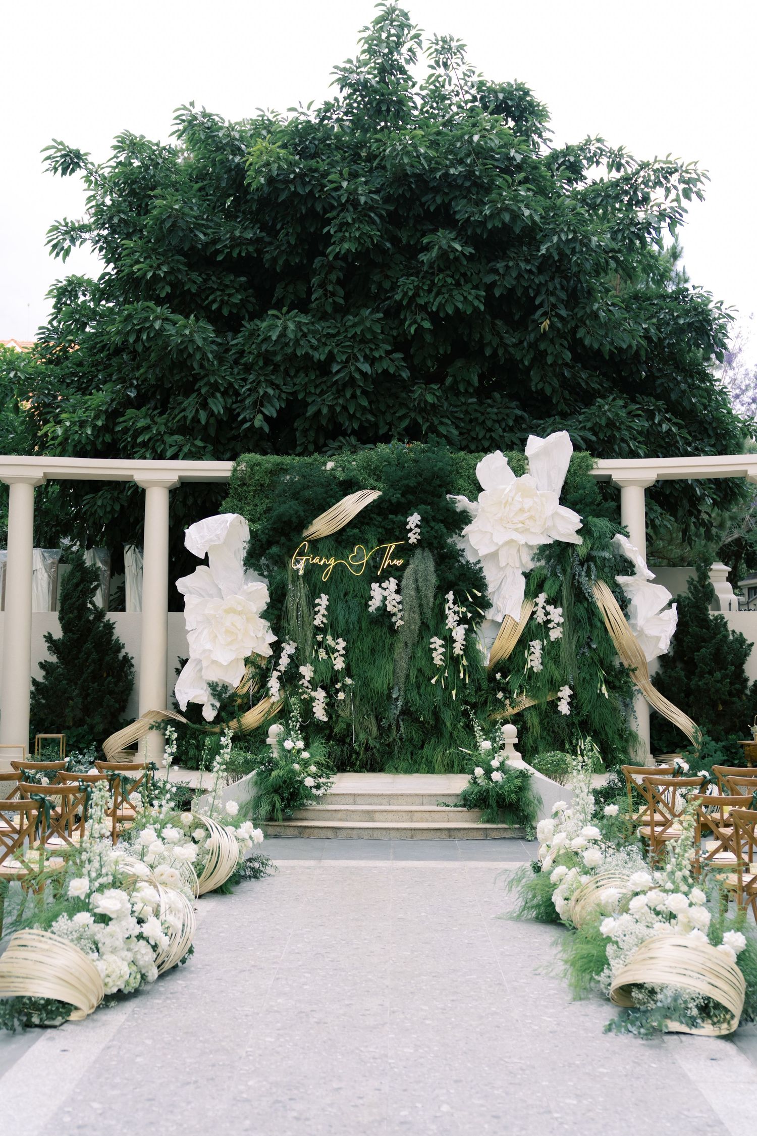 Elegant outdoor wedding ceremony aisle with lush green floral arch, white blooms, and wooden chairs.