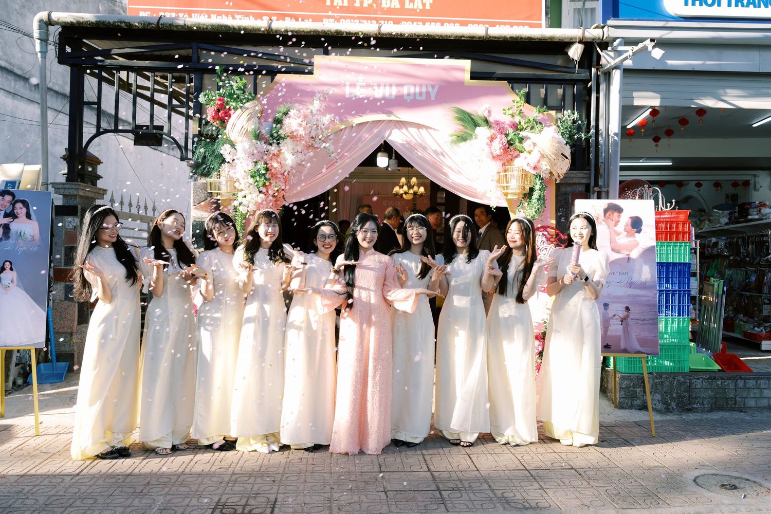 A group of women in elegant white ao dai traditional Vietnamese dresses pose together at a floral wedding arch outdoors.