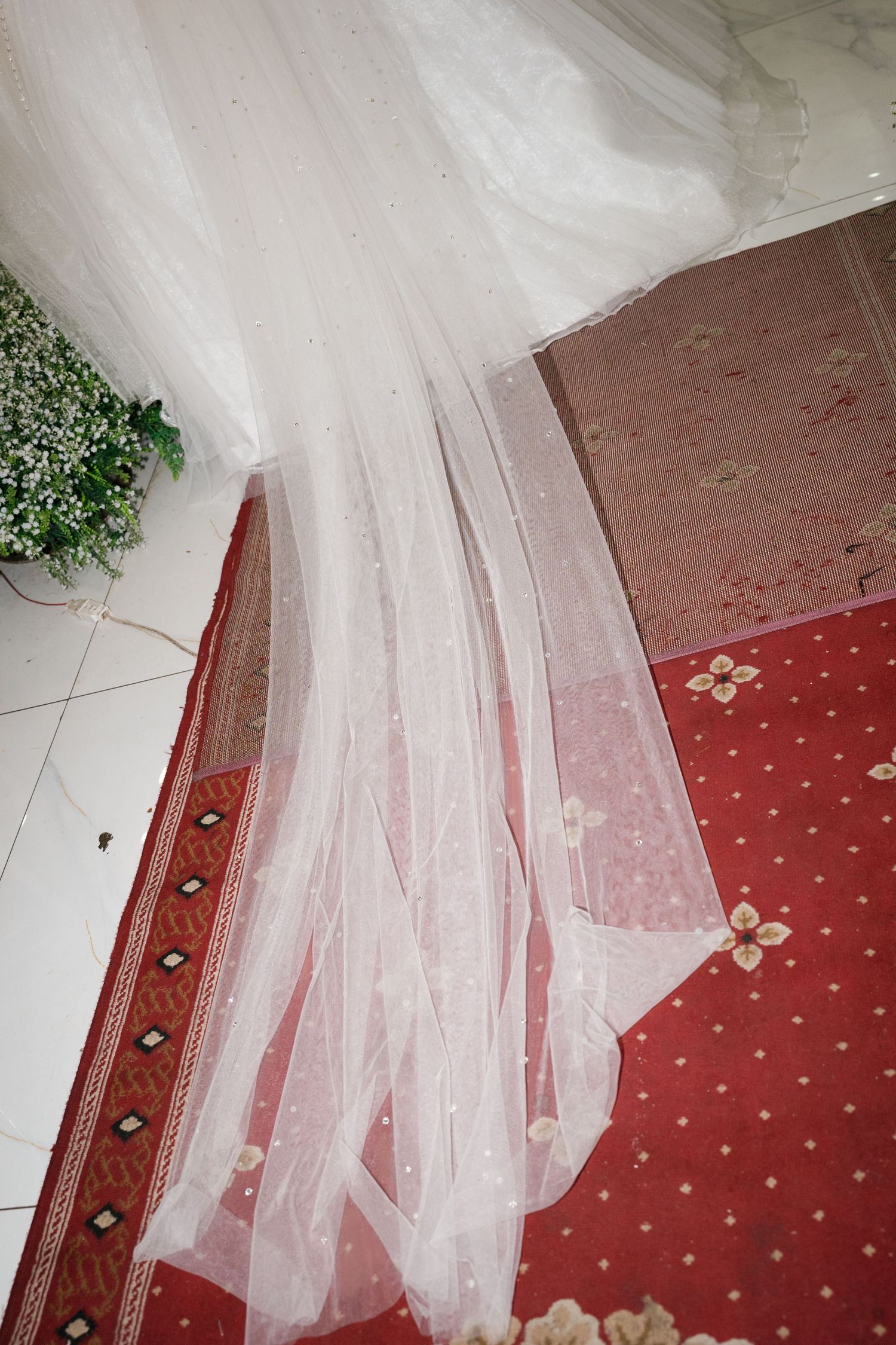 Close-up of white wedding dress veil trailing on red patterned carpet with small white flowers.