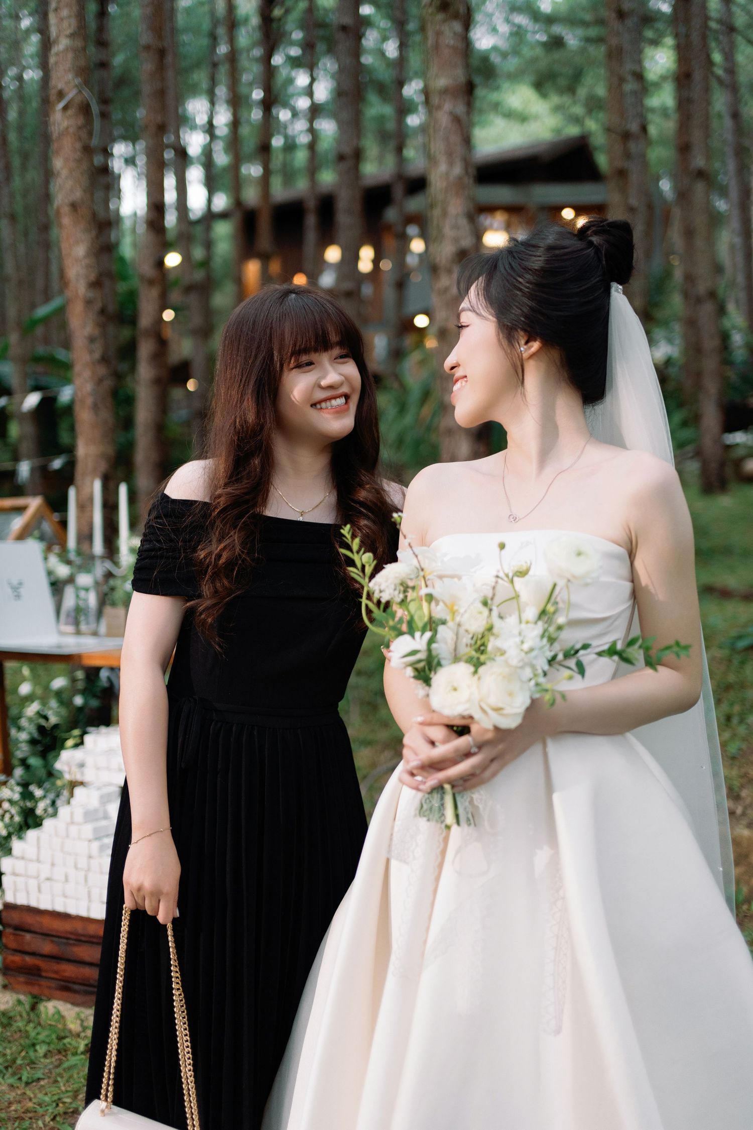 Bride in white gown holding bouquet smiling with a guest in black dress outdoors.