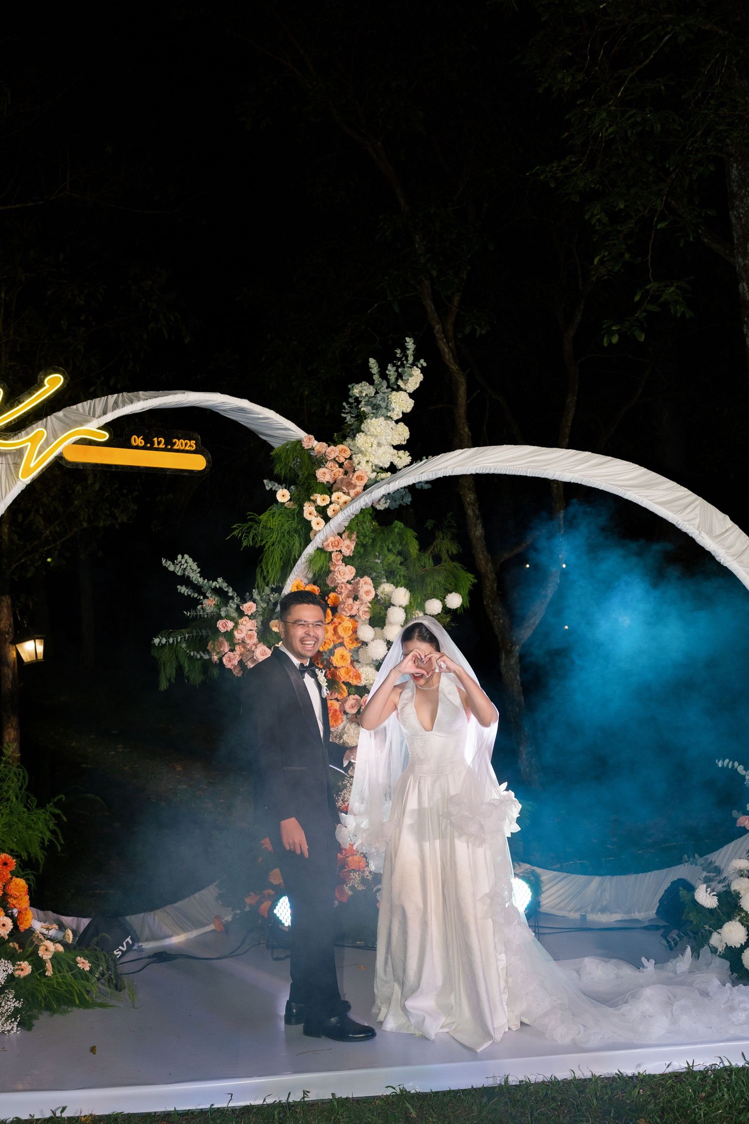 Bride and groom stand under illuminated arch with blue smoke effects at outdoor night wedding.