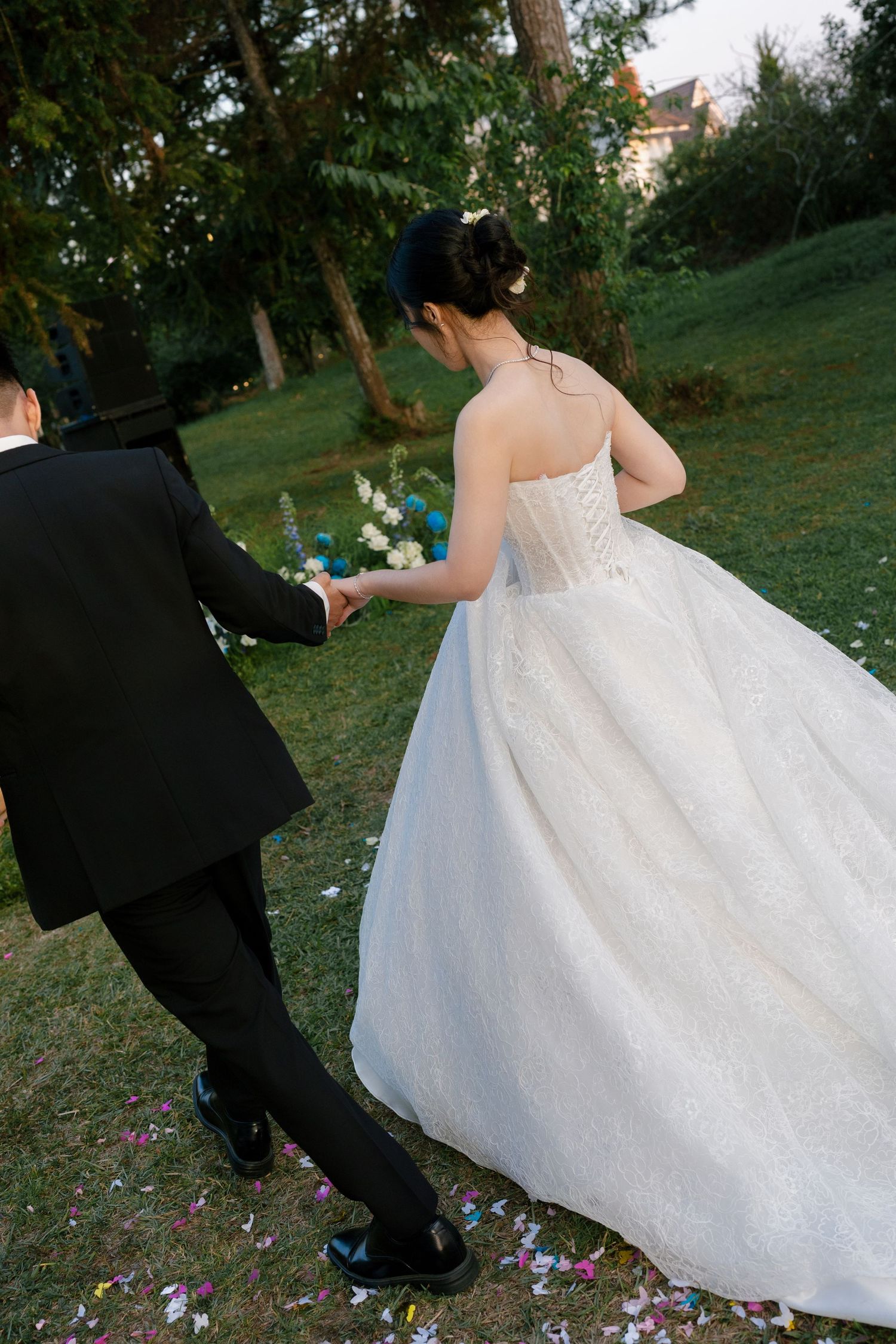 Bride in strapless white ball gown holding groom's hand walking across a petal-strewn garden path.