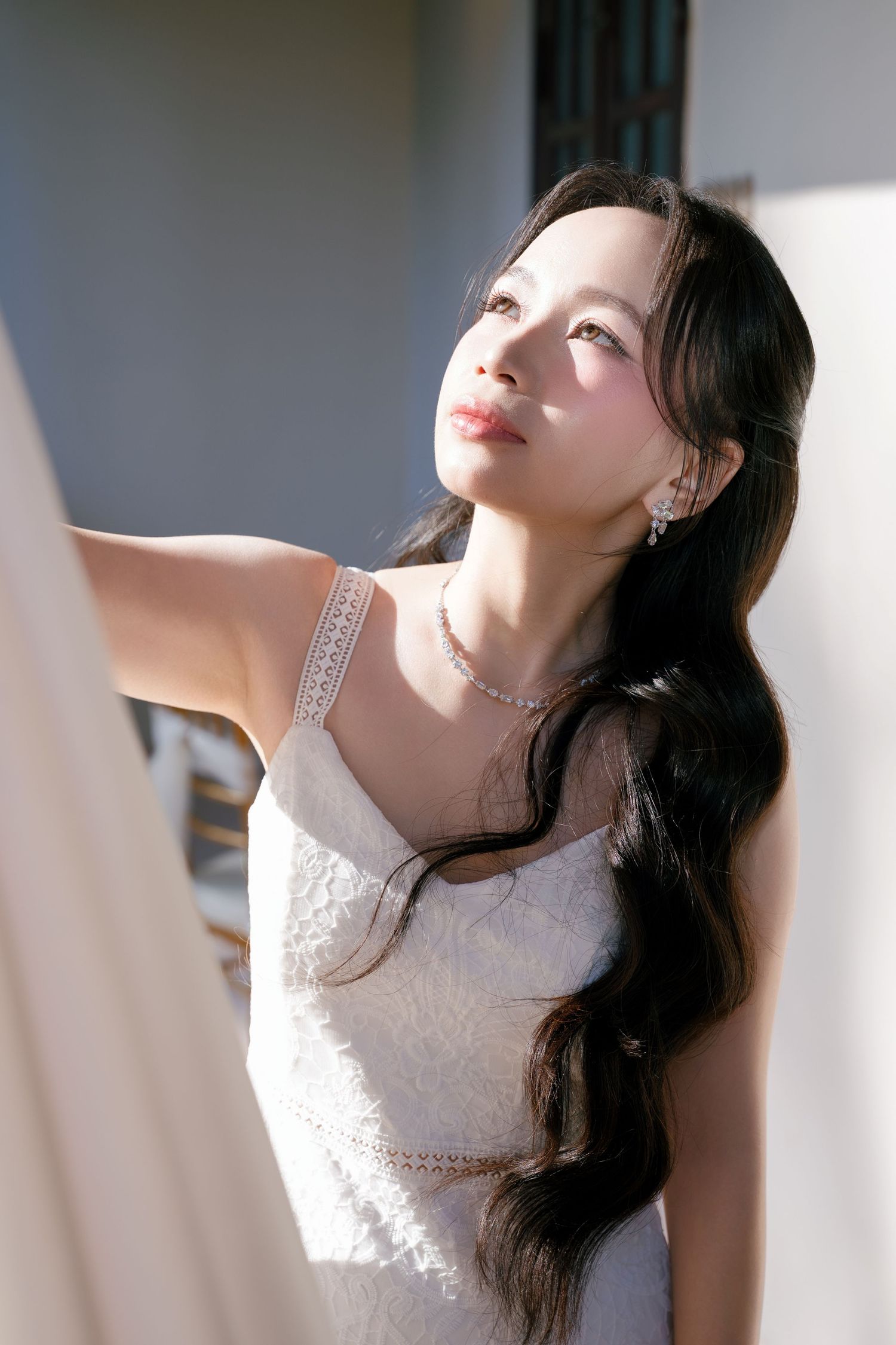 Young Asian woman in white lace dress with long wavy hair, wearing pearl jewelry, gazing upward in soft sunlight.