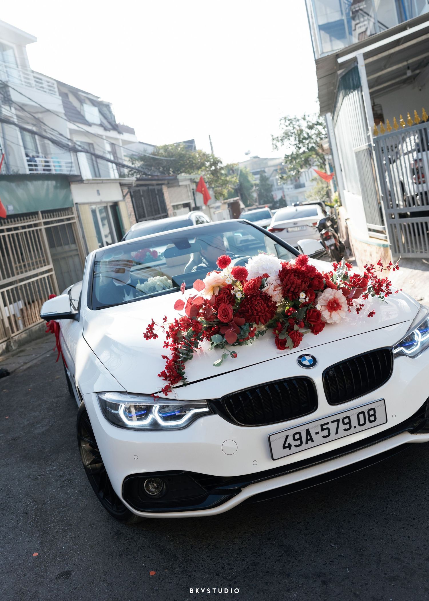 White BMW convertible adorned with red and white floral wedding decorations on the hood, parked on a street.