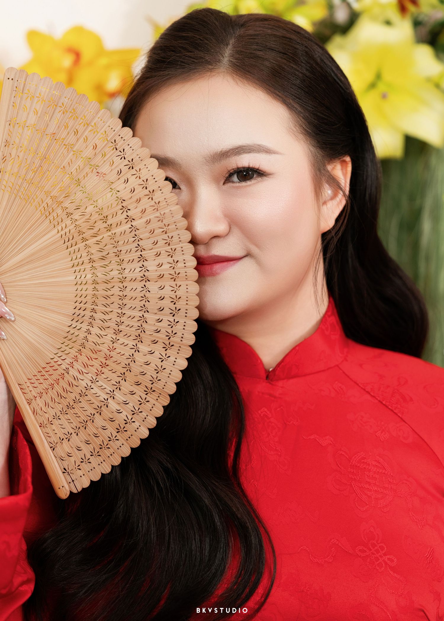 Young woman in red traditional ao dai holding a decorative wooden fan, smiling warmly with yellow flowers behind.