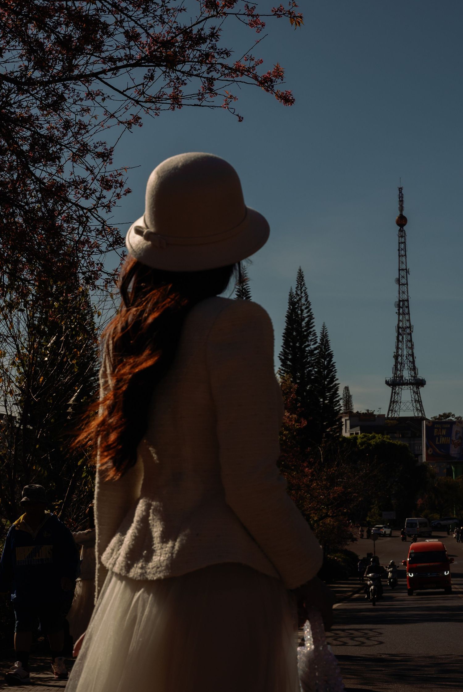 Woman in white hat and outfit stands with back turned, gazing at a tower among pine trees under a clear blue sky.