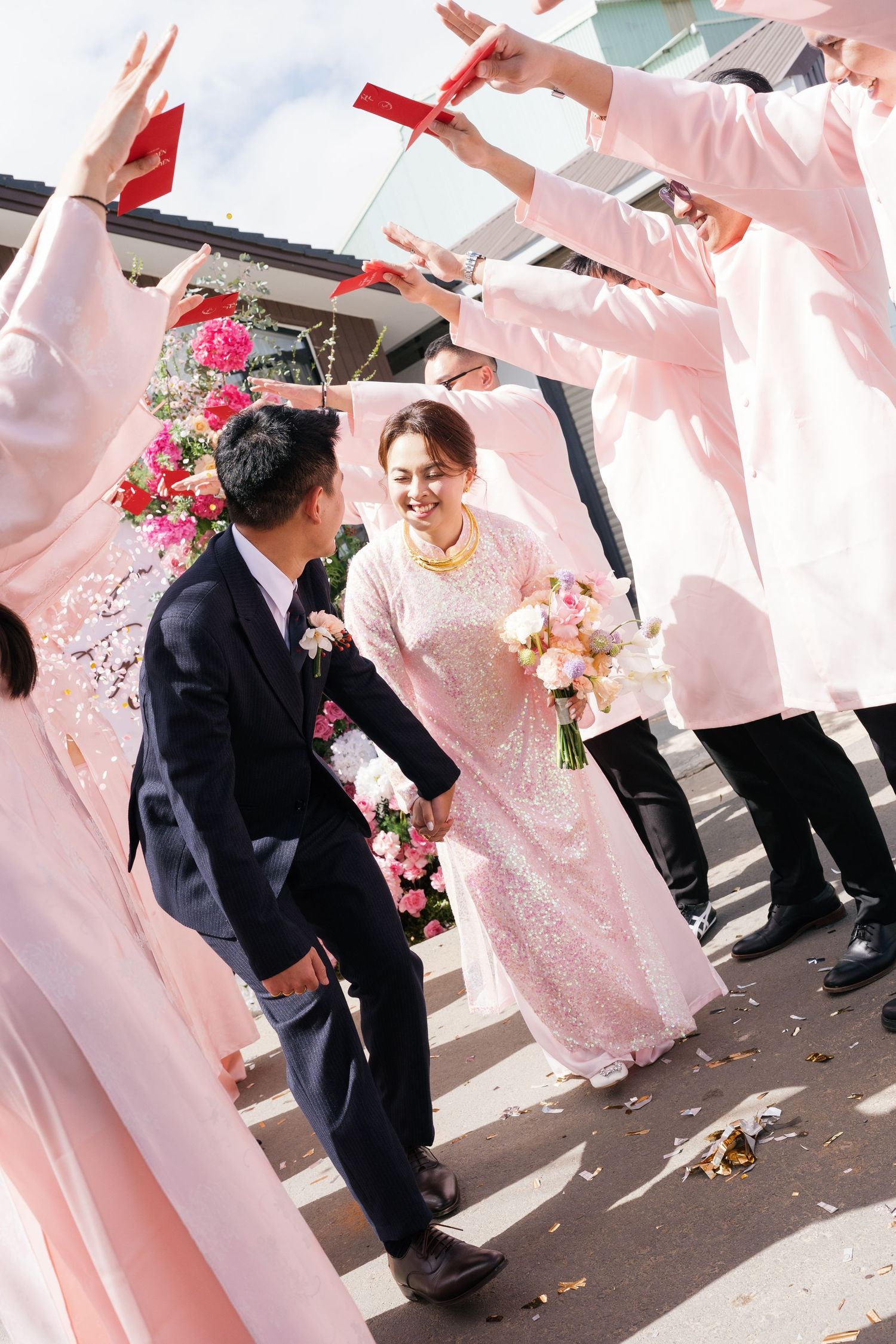 Bride and groom in traditional Vietnamese ao dai walk through arch of raised red envelopes held by wedding party outdoors.