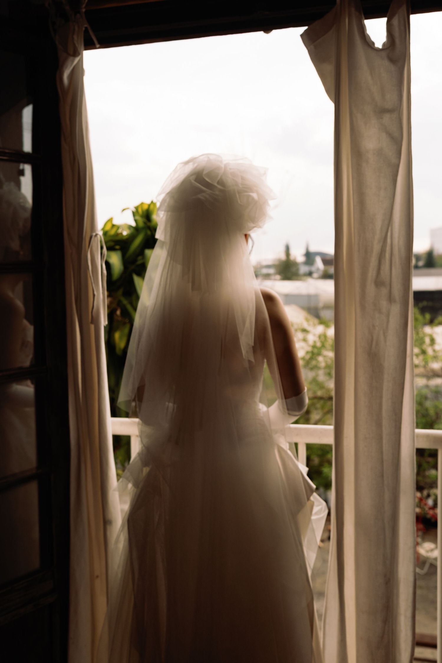 Bride in white wedding dress and veil stands at open balcony door, gazing at outdoor scenery on cloudy day.