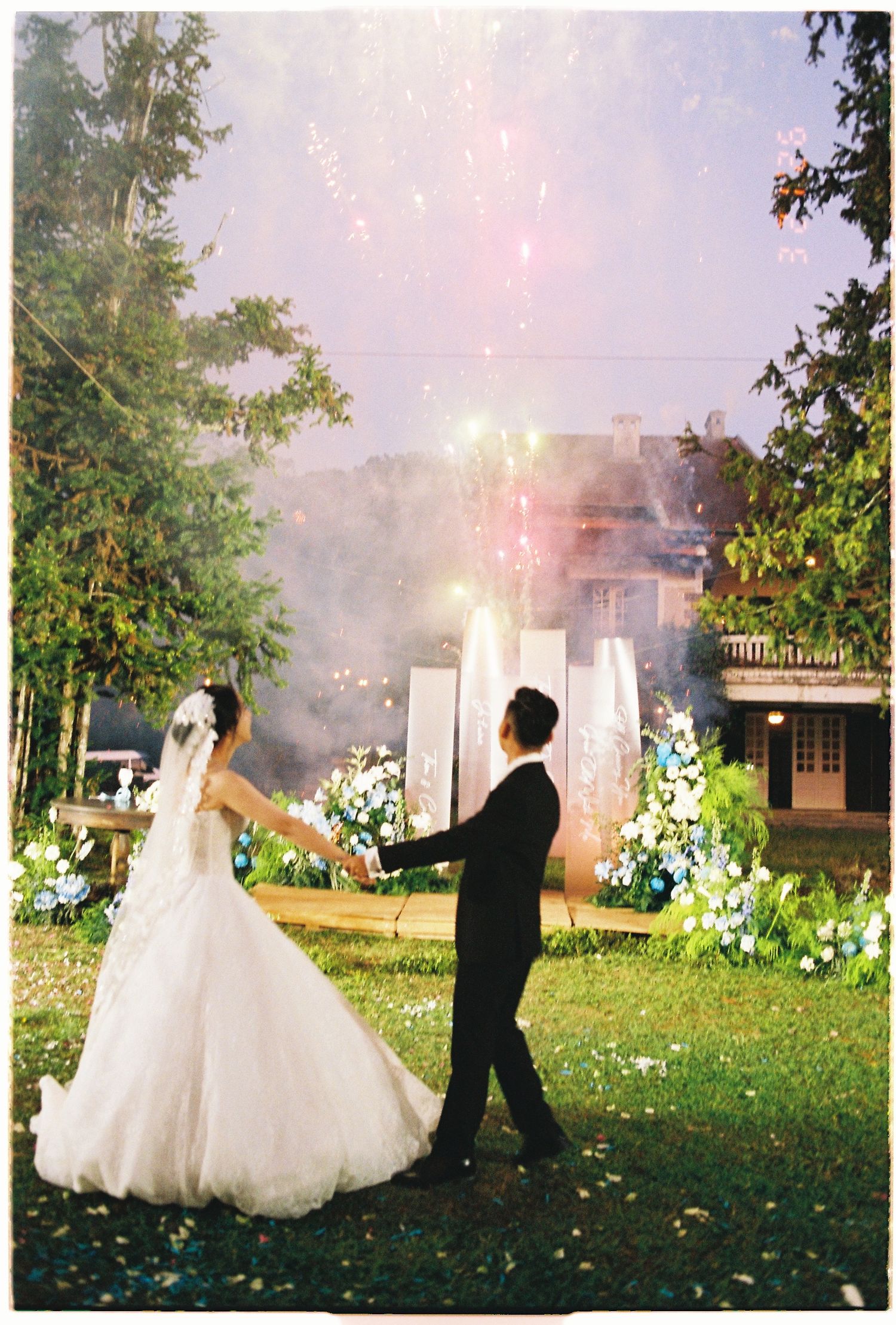 Bride and groom holding hands at outdoor wedding with fireworks and floral arch backdrop at dusk.