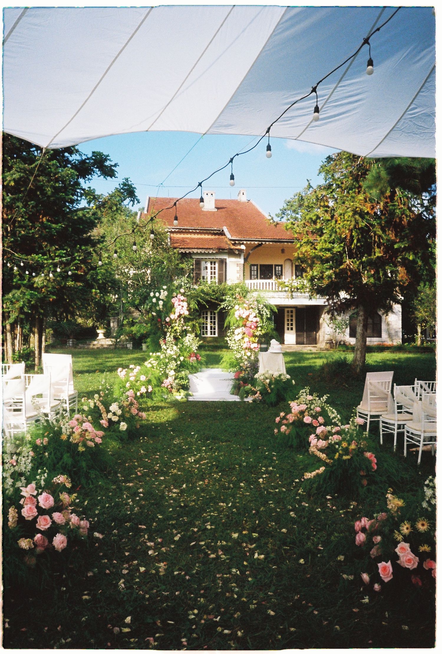 Elegant outdoor wedding ceremony setup with pink floral arrangements, white chairs, and string lights in a lush garden estate.