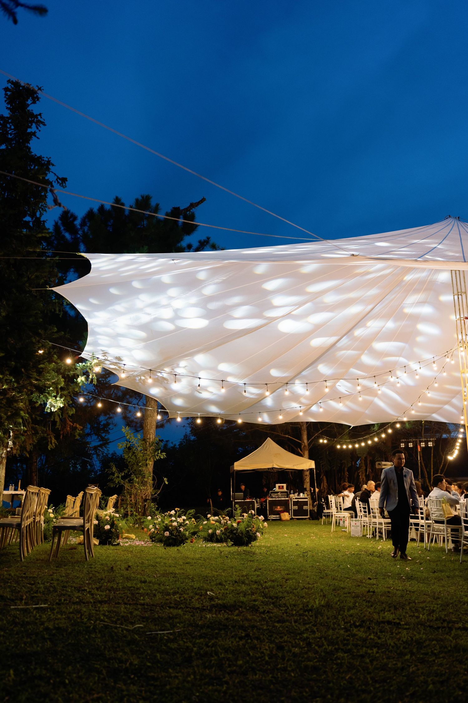 Outdoor evening wedding reception under illuminated white canopy with string lights on lush green lawn at twilight.