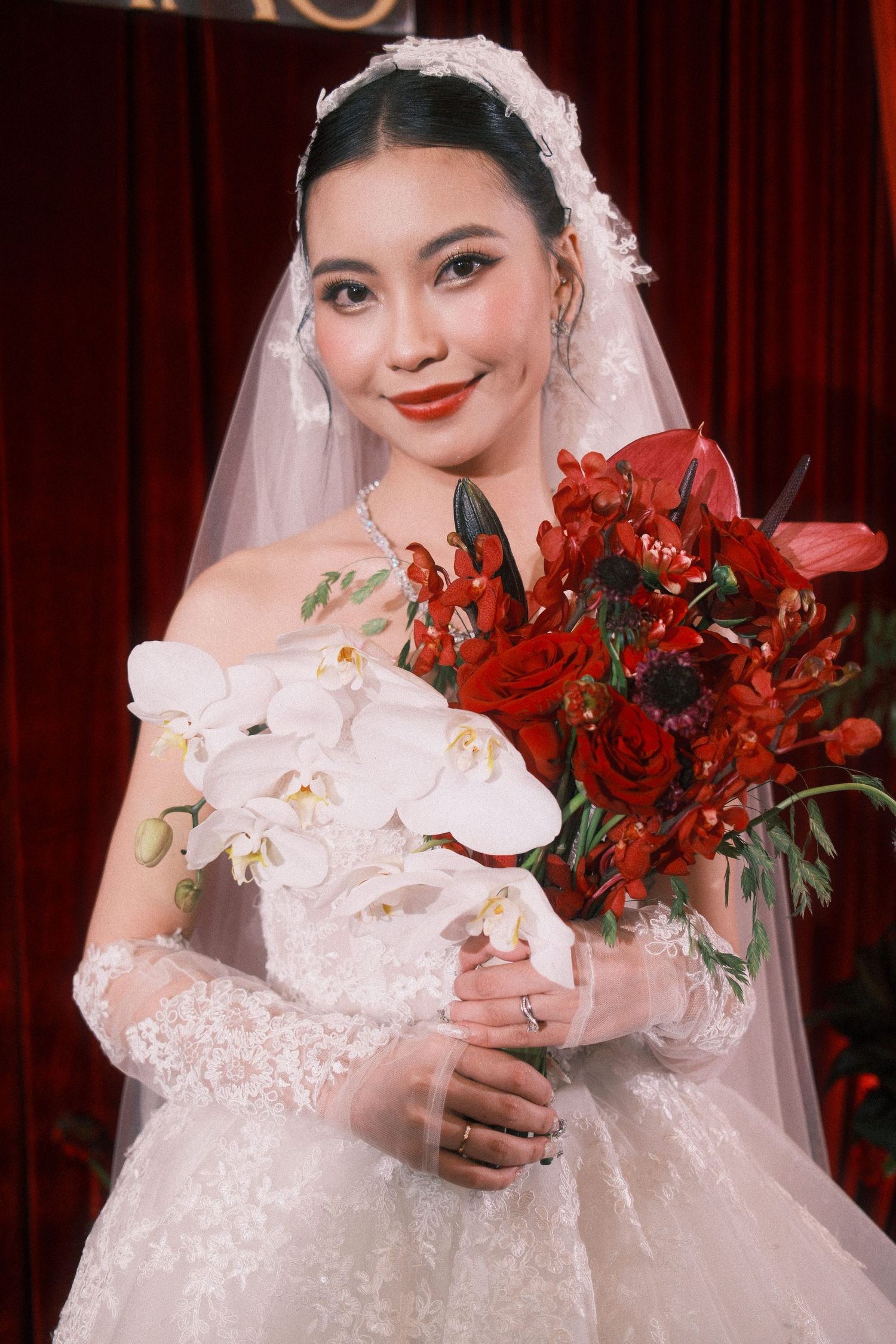 A bride in a white lace gown and veil holds a red and white floral bouquet, smiling elegantly.