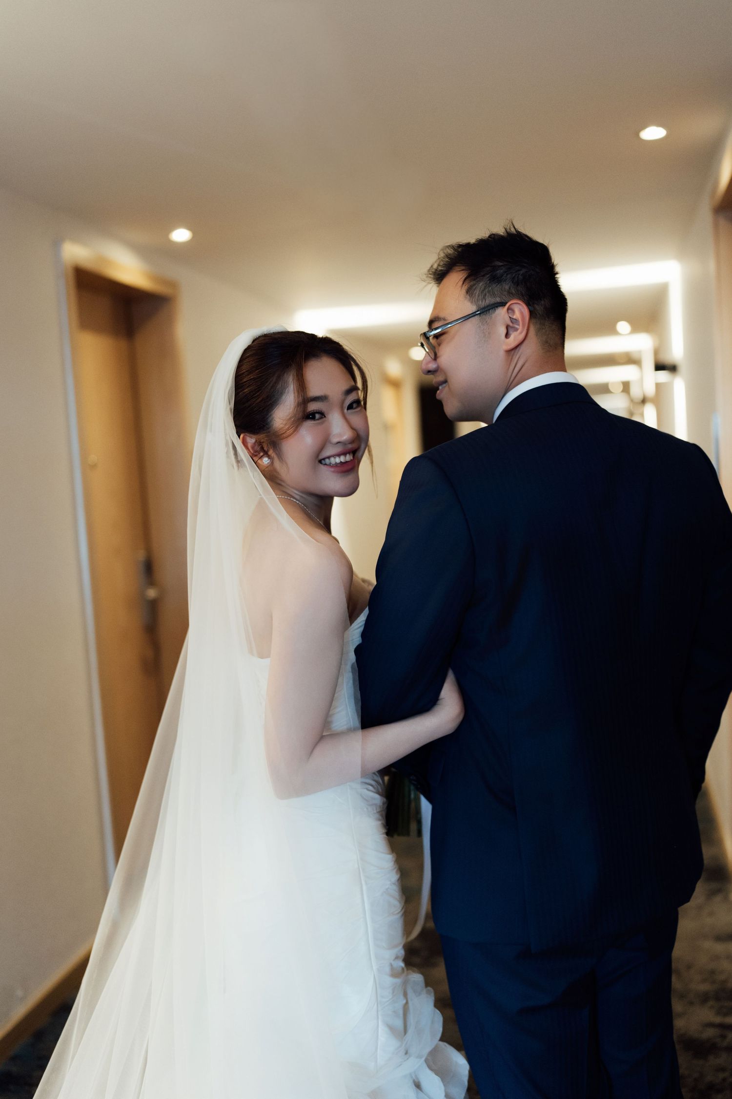 Smiling Asian bride in white wedding gown and veil gazes back while embracing groom in navy suit in hotel hallway.