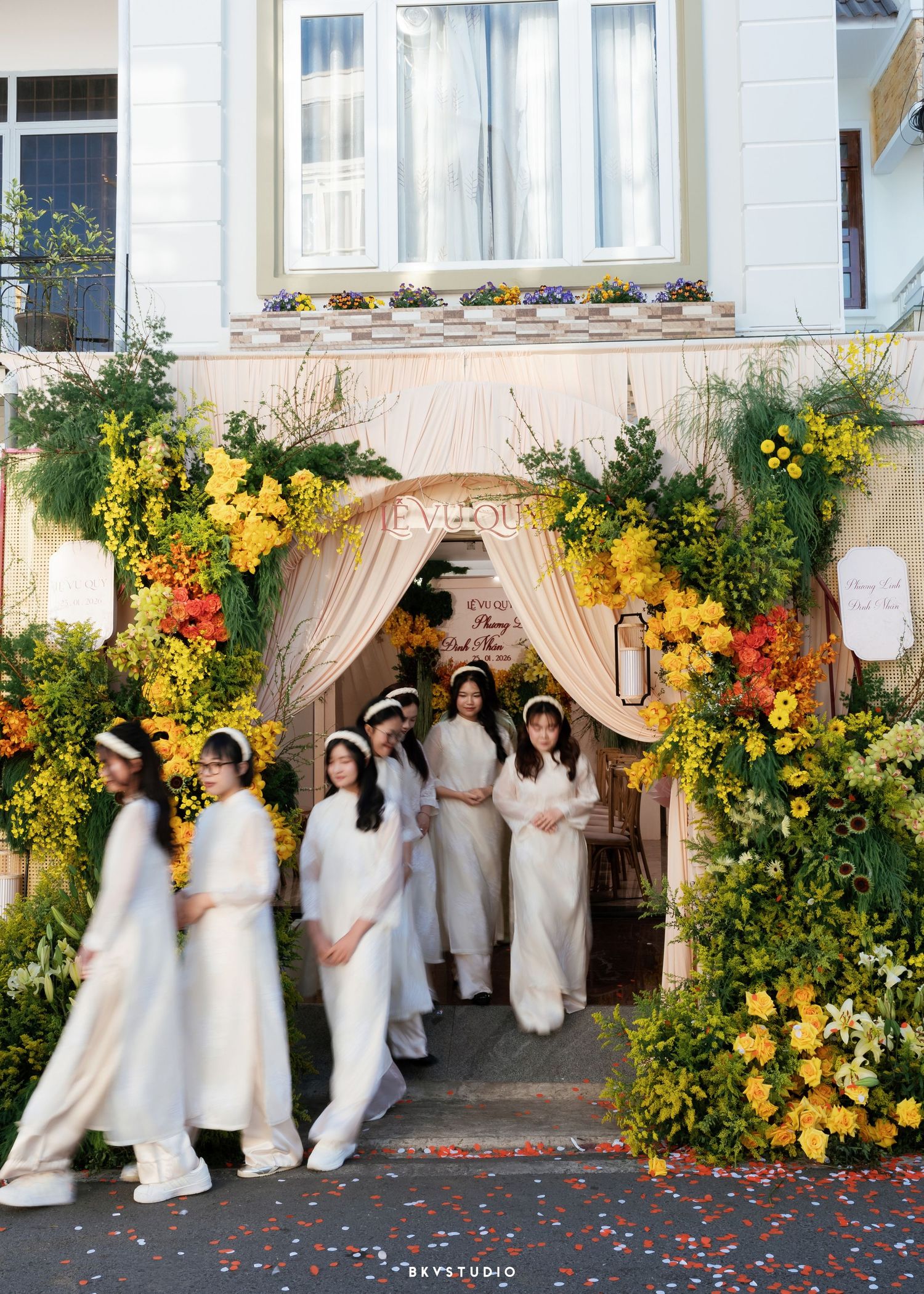 Women in white ao dai gowns walk through an elegant floral arch adorned with yellow and green blooms at a Vietnamese wedding.