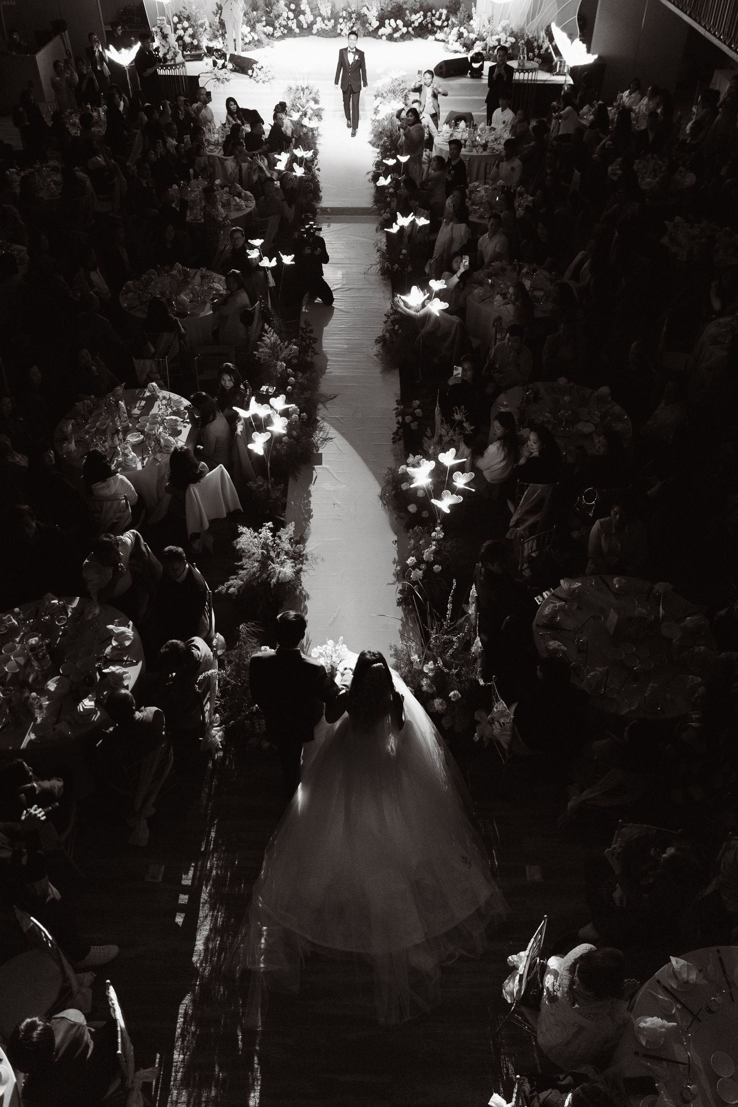 Aerial black and white photo of bride walking down aisle at grand wedding ceremony with guests.
