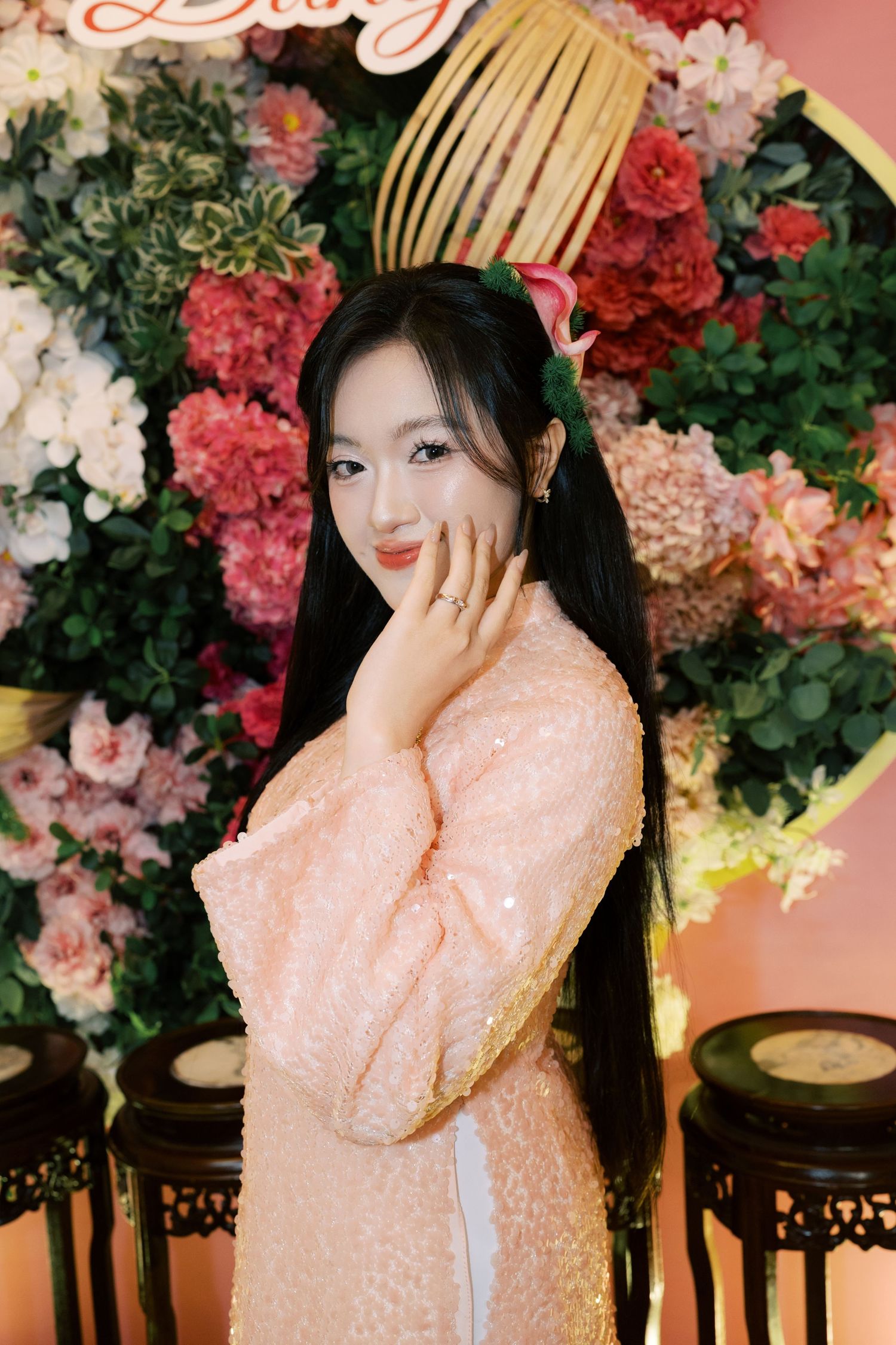 Young woman in pink sequin ao dai poses gracefully before a vibrant floral backdrop with red and white blooms.