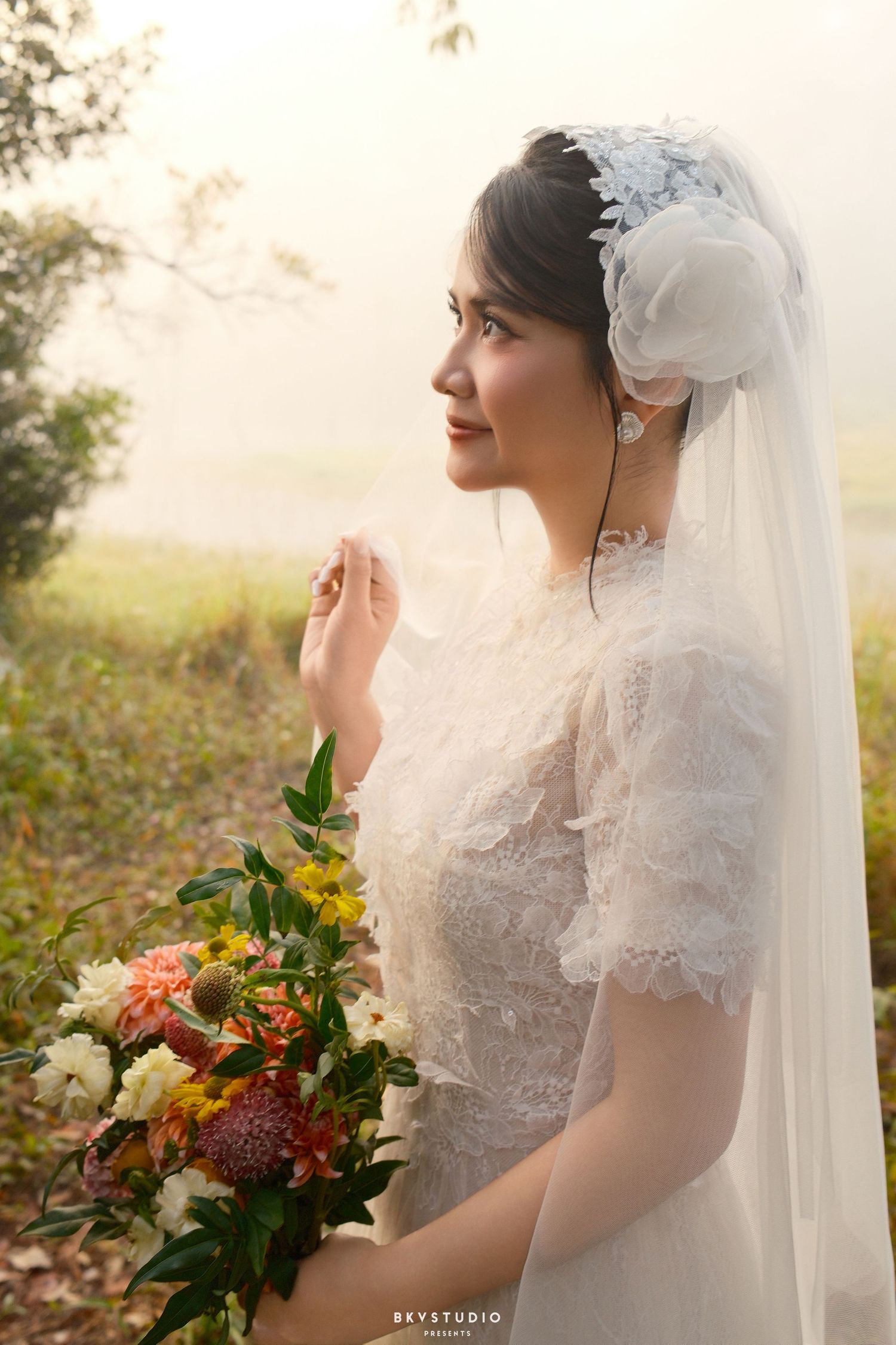 A bride in a white lace dress and veil holds a colorful bouquet in a misty outdoor setting.