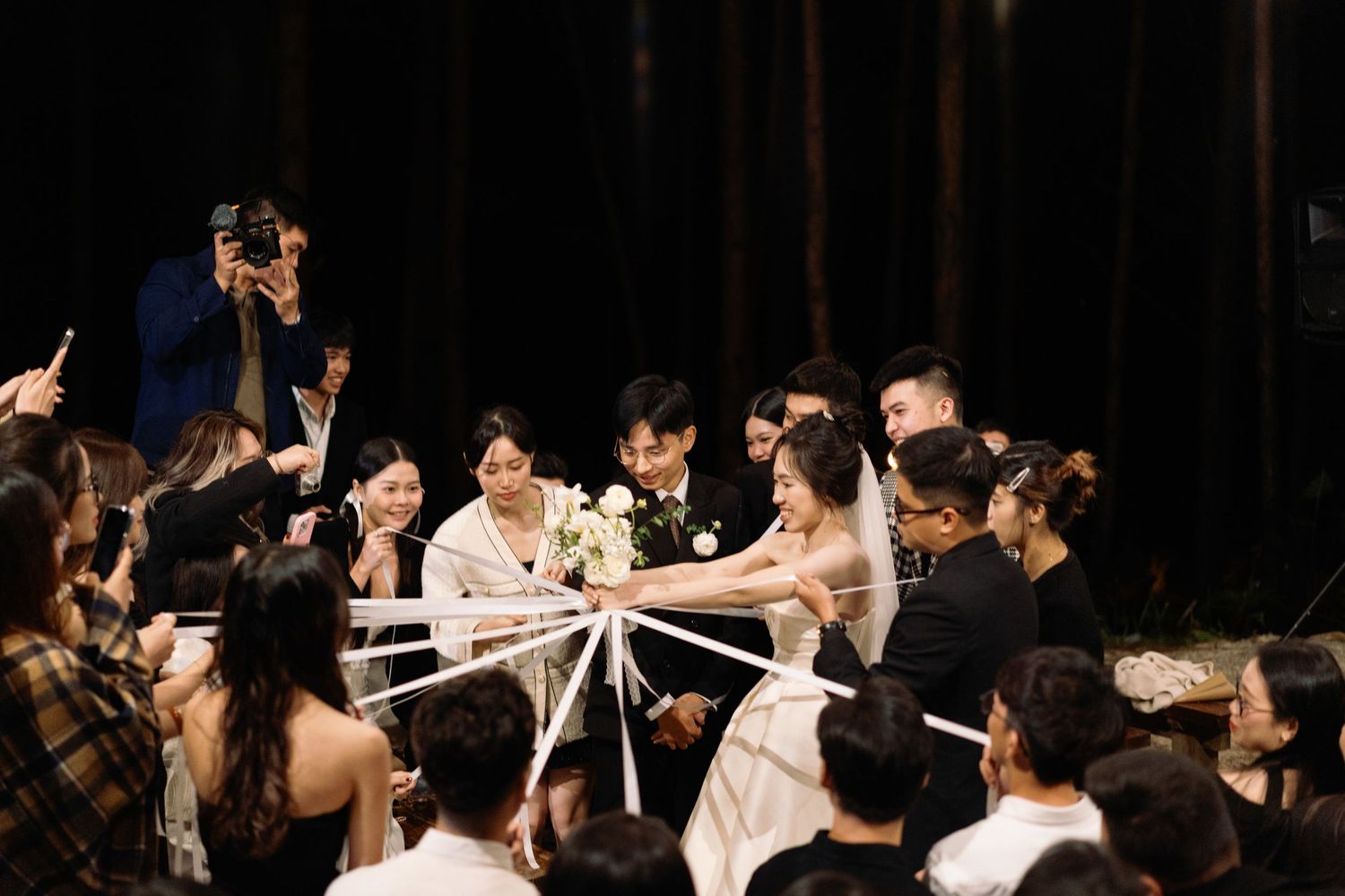Bride and groom surrounded by guests holding ribbons during a unity ceremony at an outdoor wedding.