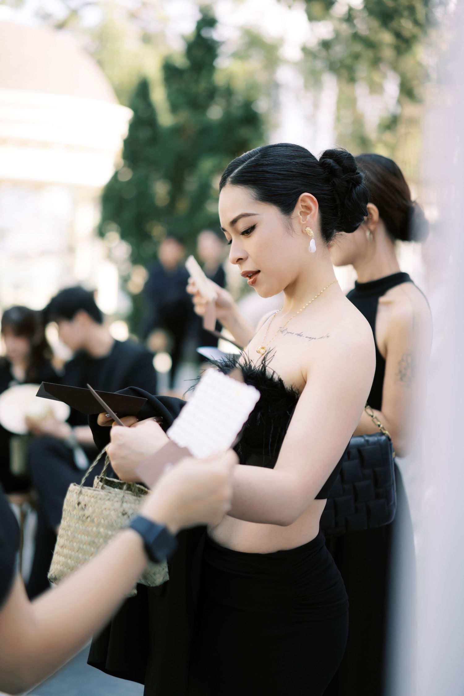 Elegant woman in black feather strapless top reads wedding program at outdoor ceremony.