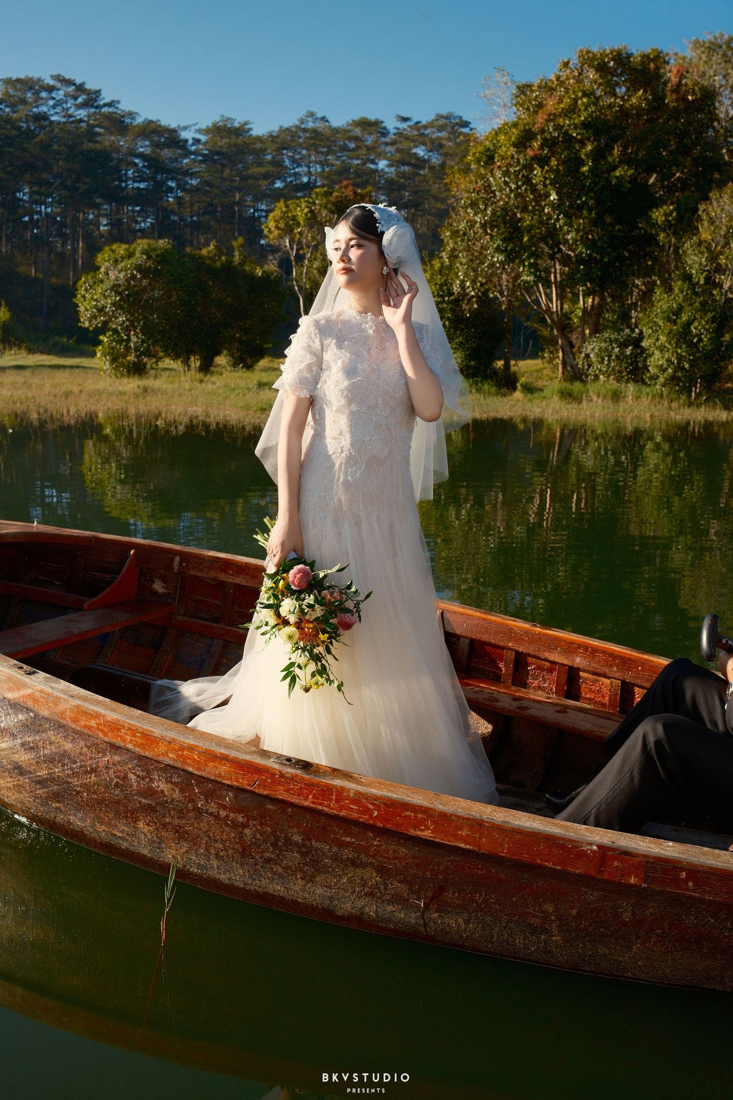 Elegant bride in feathered wedding gown stands in wooden boat holding bouquet on sunny lakeside.