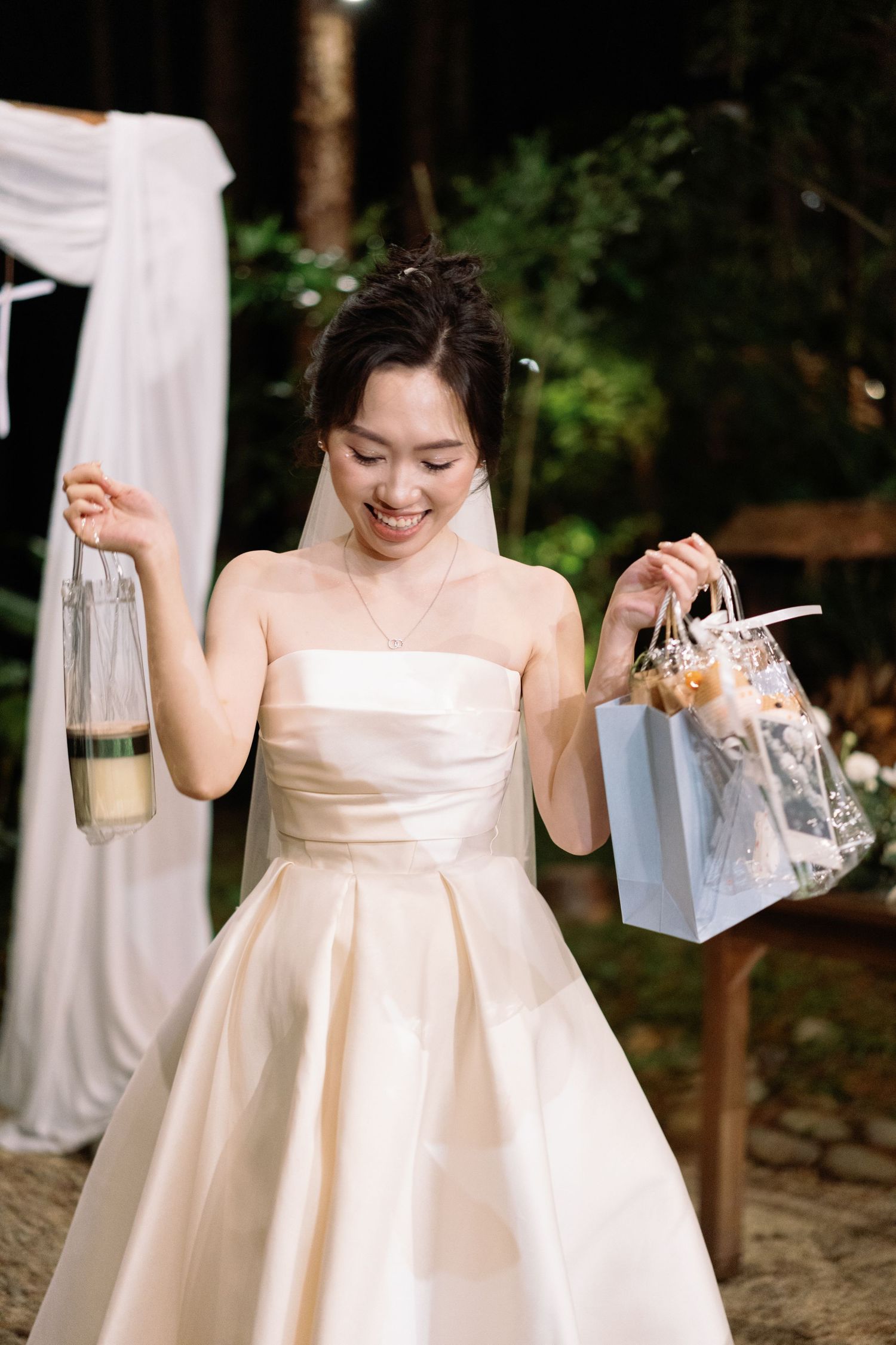 Smiling bride in strapless gown holding a lantern and gift bag at an outdoor forest wedding.