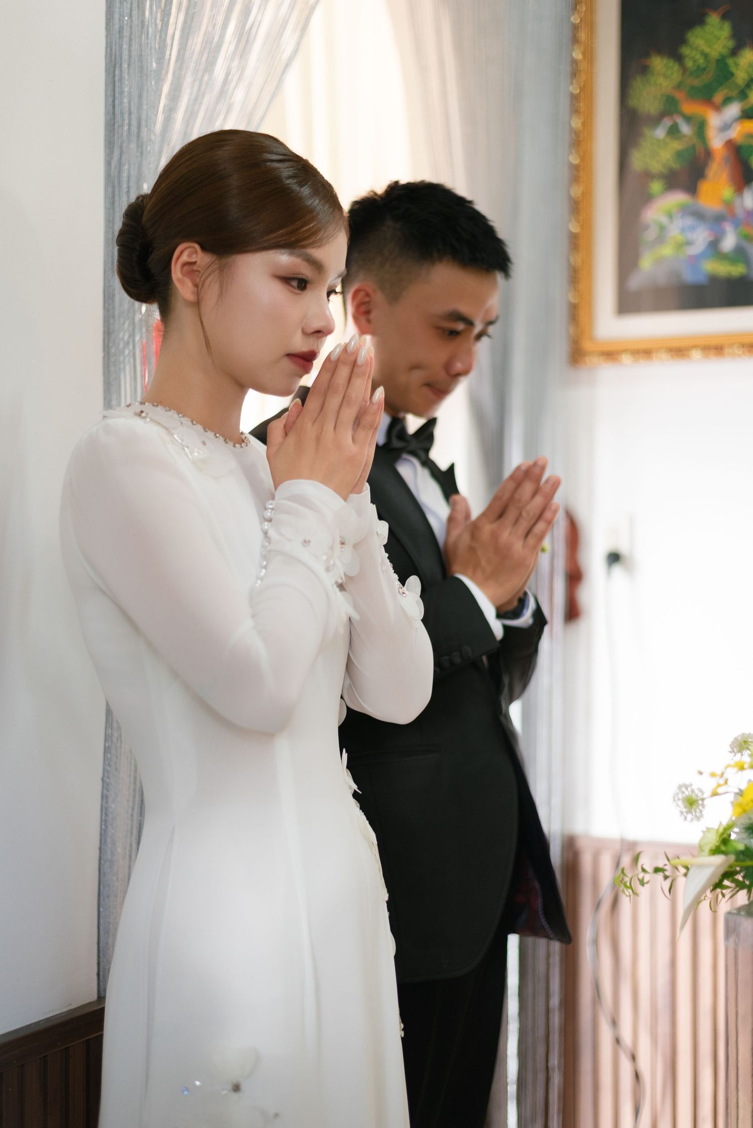 Bride in white ao dai and groom in tuxedo praying together during a traditional Vietnamese wedding ceremony.