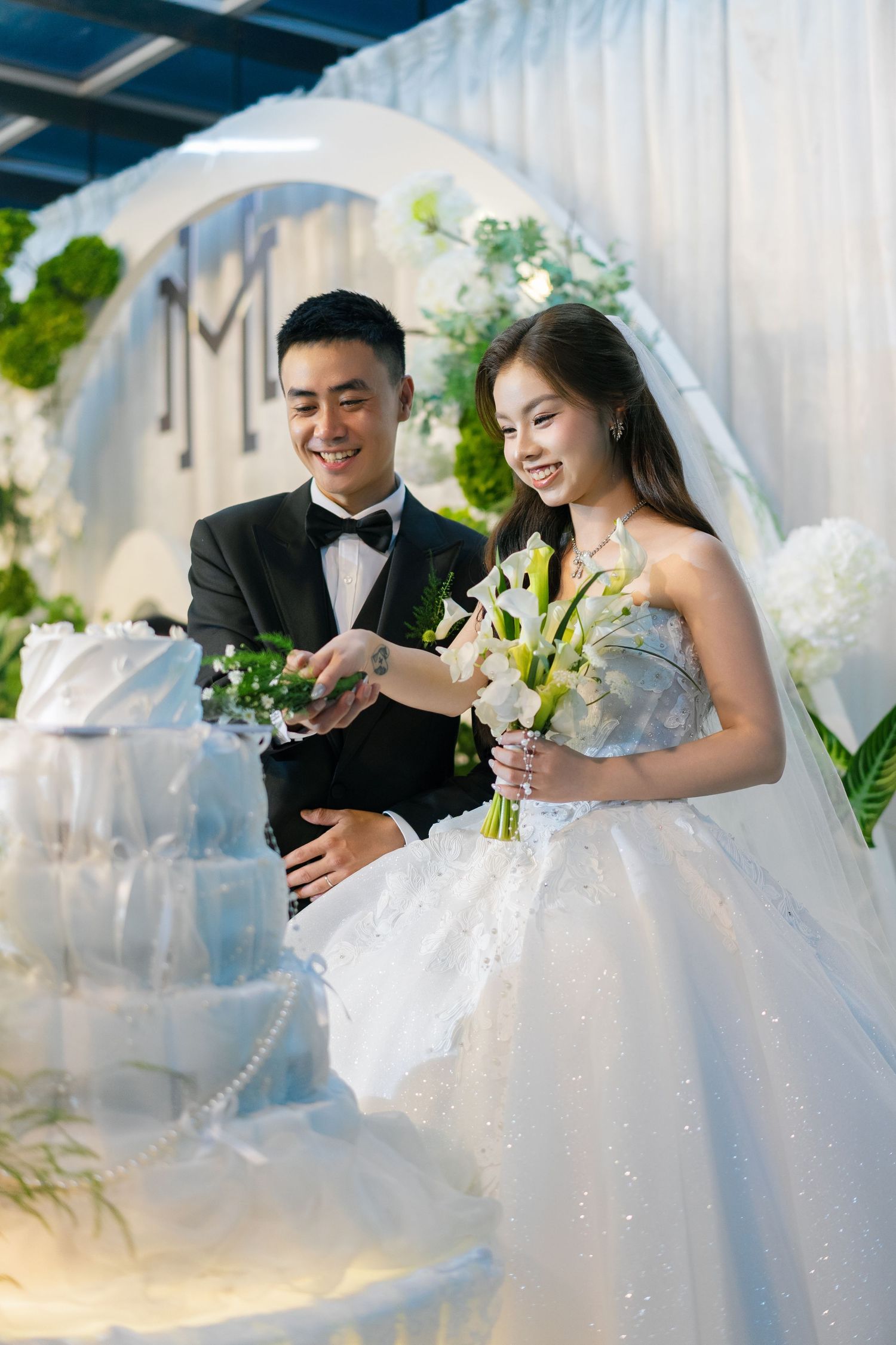 Asian bride and groom smiling while cutting their elegant white tiered wedding cake at a floral decorated venue.