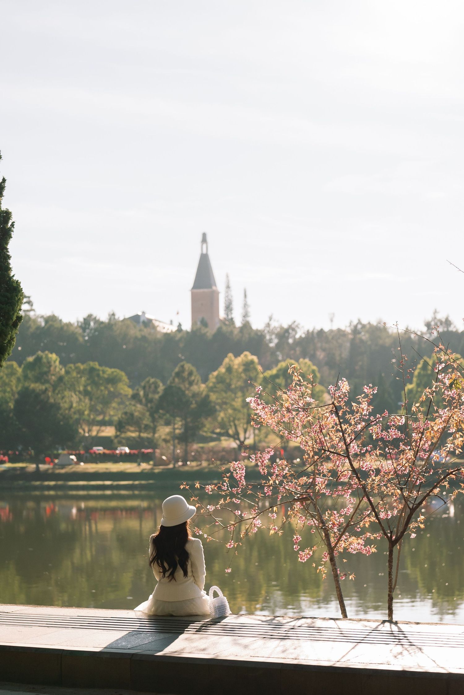 Woman in white dress and hat sitting by a tranquil lake with cherry blossoms and a church tower in the background.