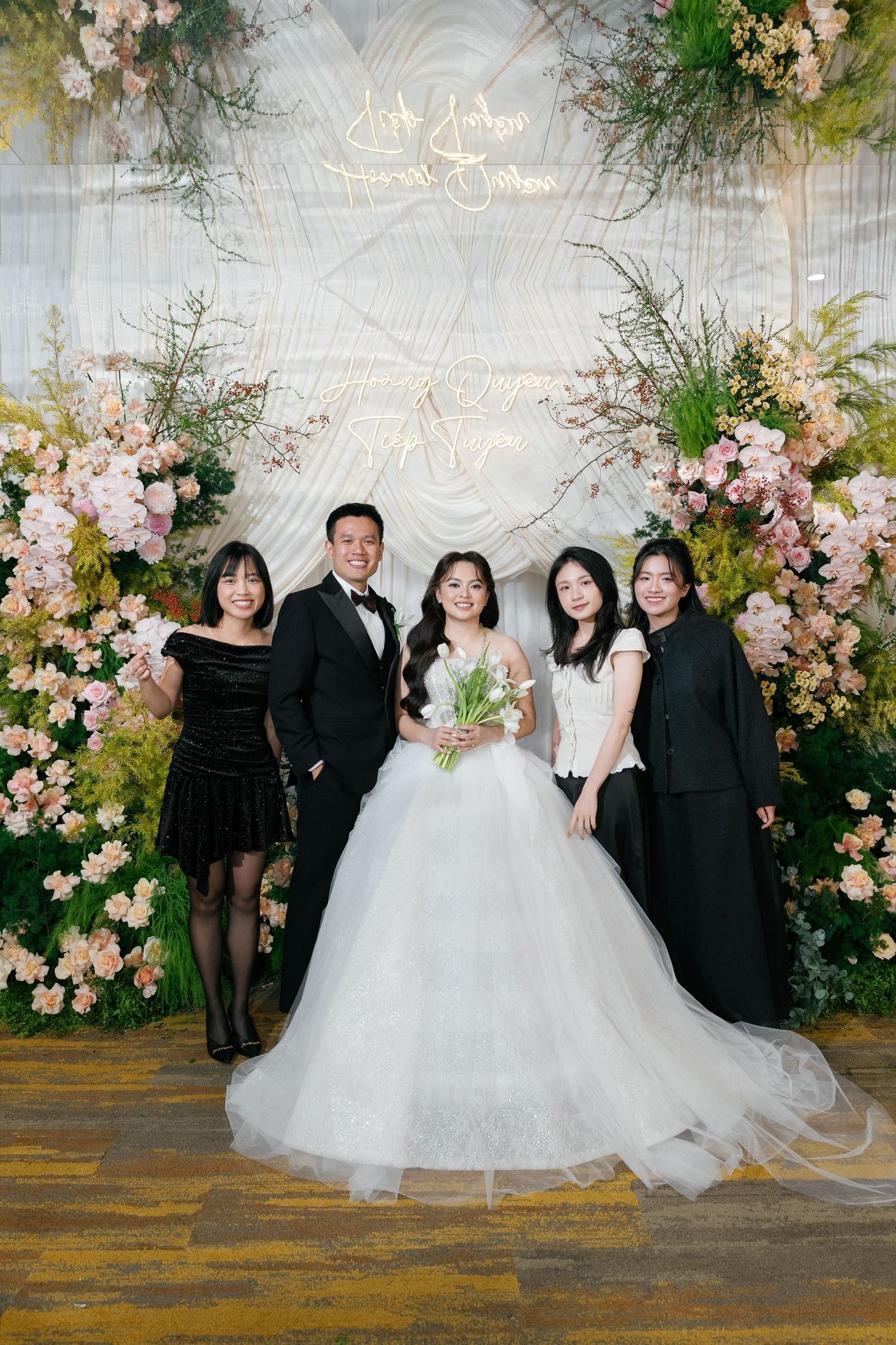 Bride in white ball gown poses with groom and guests before a floral wedding backdrop with pink blooms.
