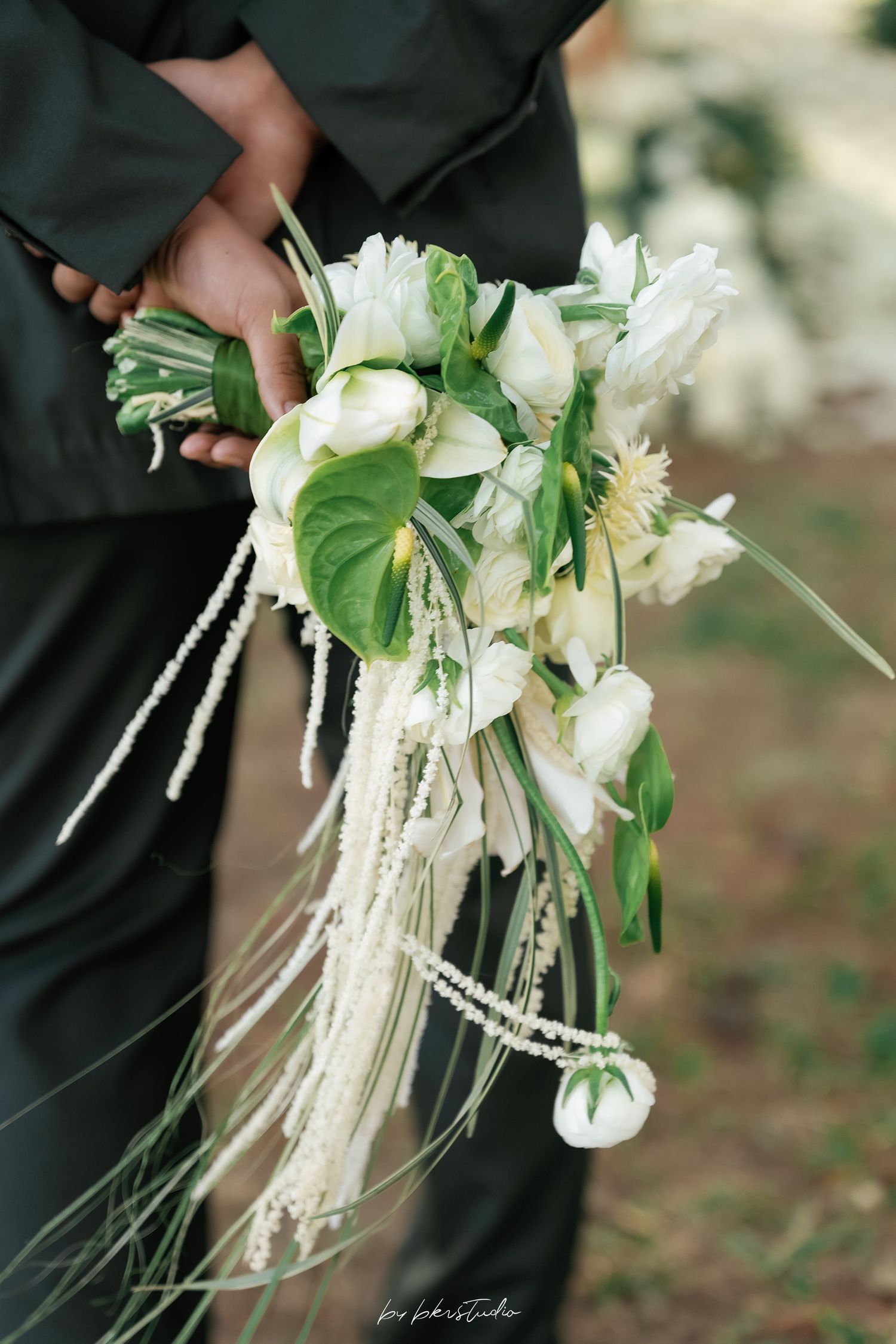 Elegant cascading bridal bouquet with white tulips, anthurium, and pearl strands held by person in black suit.