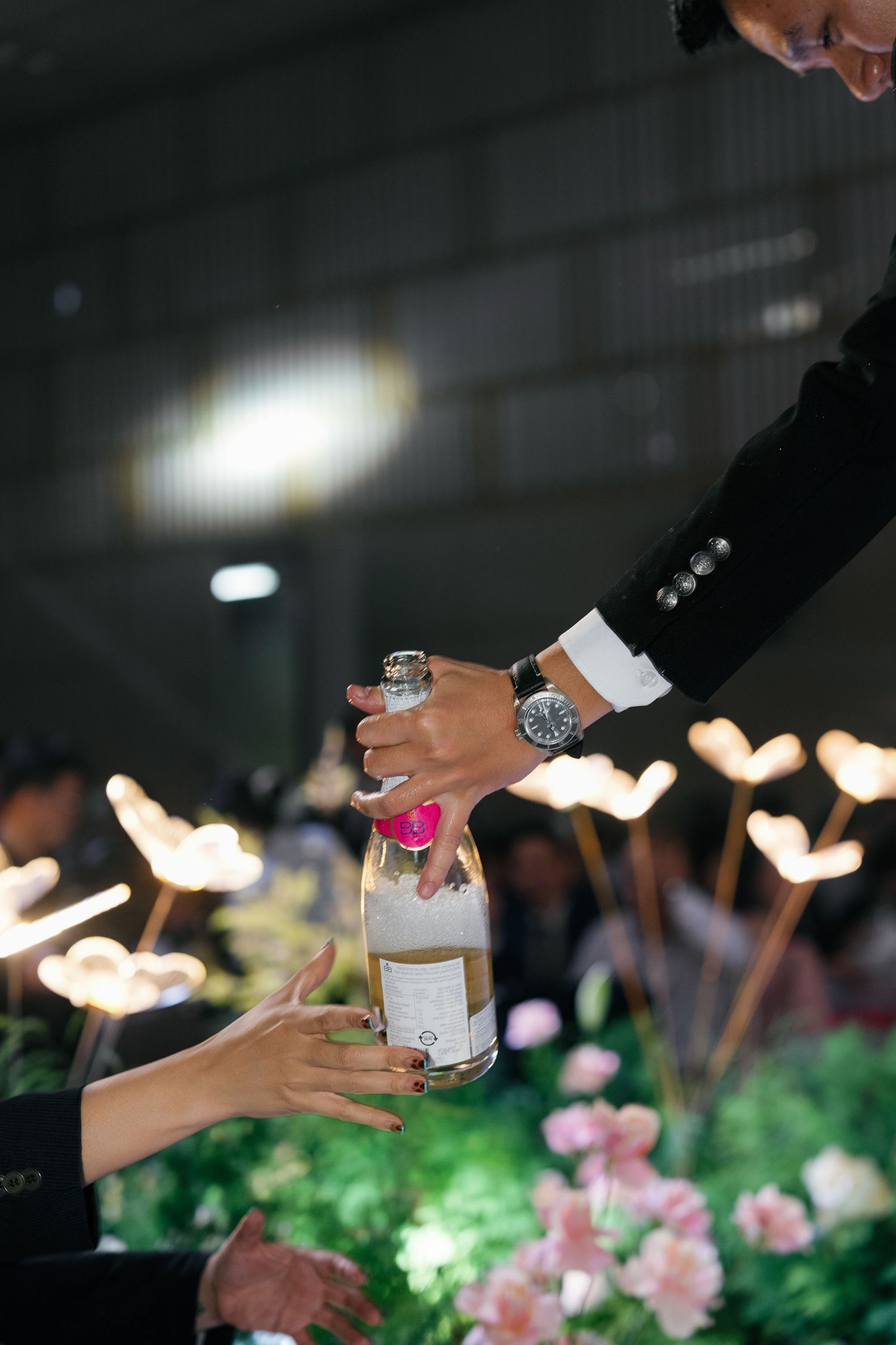 Person handing a bottle of champagne over floral arrangements with heart-shaped lights at an elegant event.