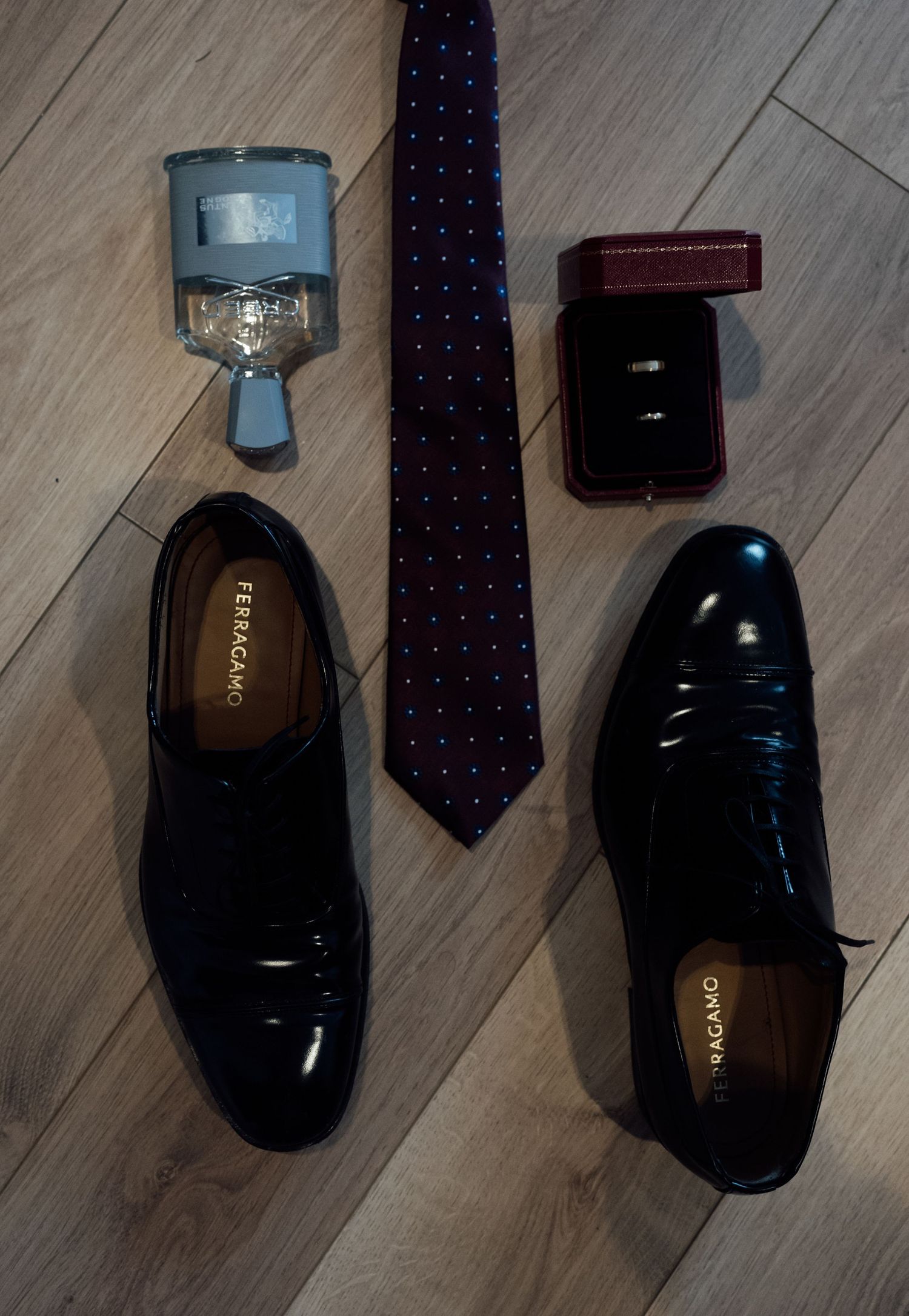 Ferragamo black patent leather dress shoes with a navy tie, cologne, and wedding rings on wood floor.