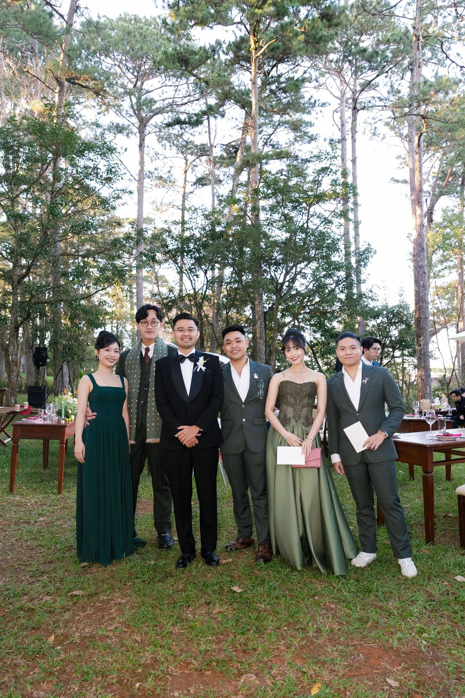 Group of wedding guests posing in green and formal attire among tall pine trees at outdoor reception.
