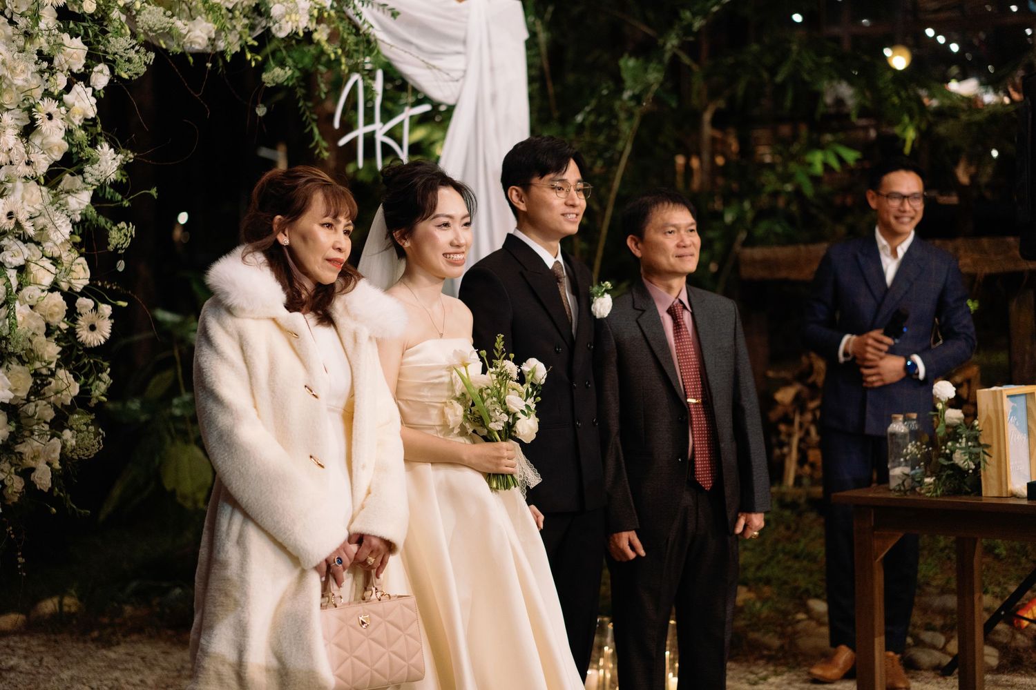 Bride and groom pose with family members at elegant outdoor wedding reception with floral arch.