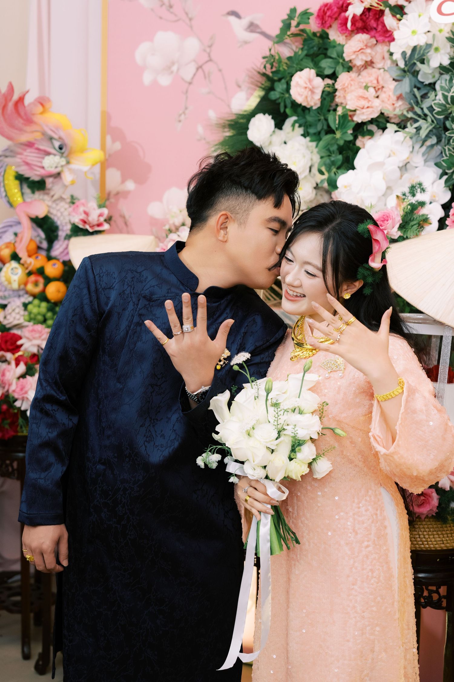 Vietnamese groom in navy ao dai kisses bride in pink dress while both display wedding rings.