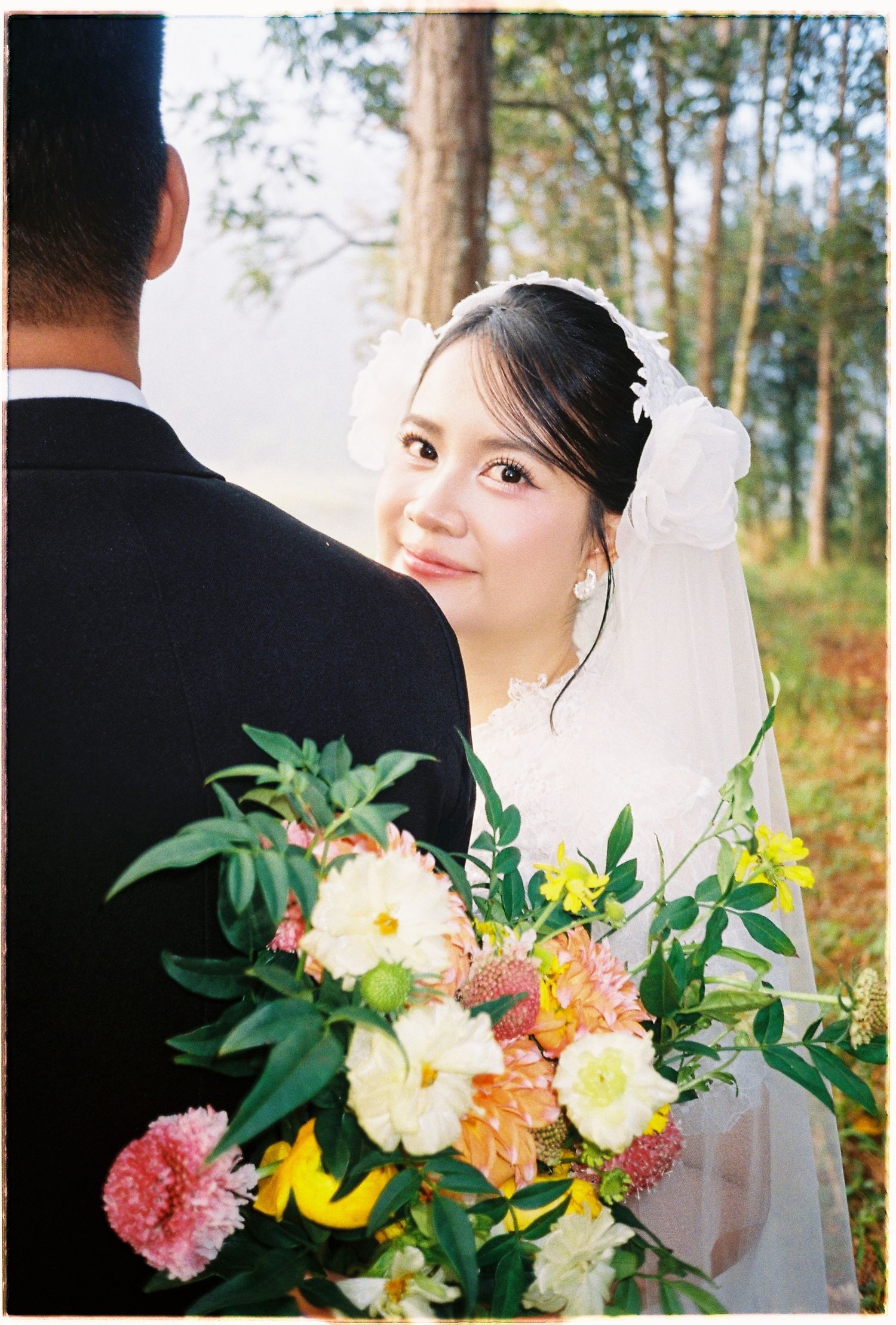 Smiling bride in white lace gown and veil looks back over shoulder holding a vibrant floral bouquet.