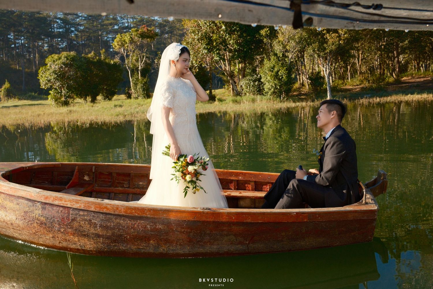 A bride in a white dress and groom sit together in a rustic wooden boat on a green lake.