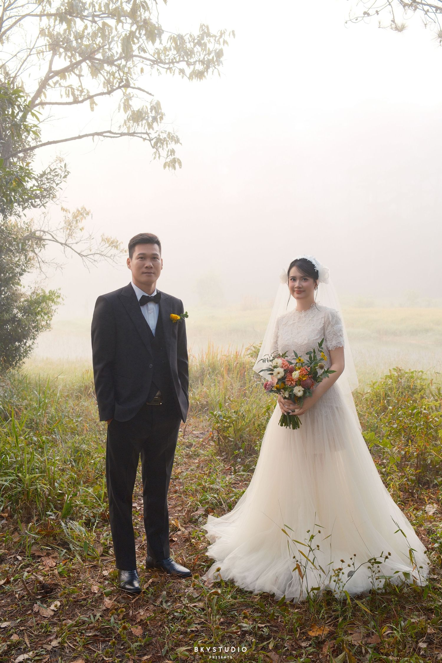 Couple poses in misty outdoor wedding shoot, groom in black tuxedo, bride in white lace gown holding colorful bouquet.