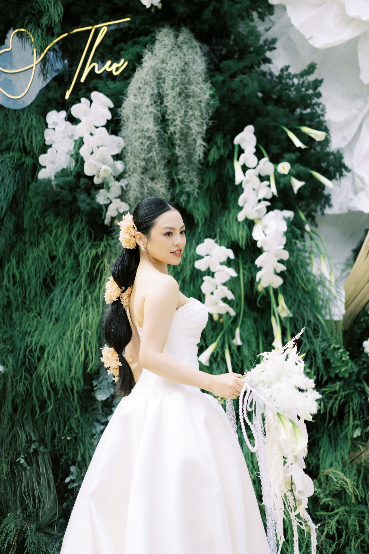 Bride in strapless white ball gown holds white bouquet before lush green floral wedding backdrop.