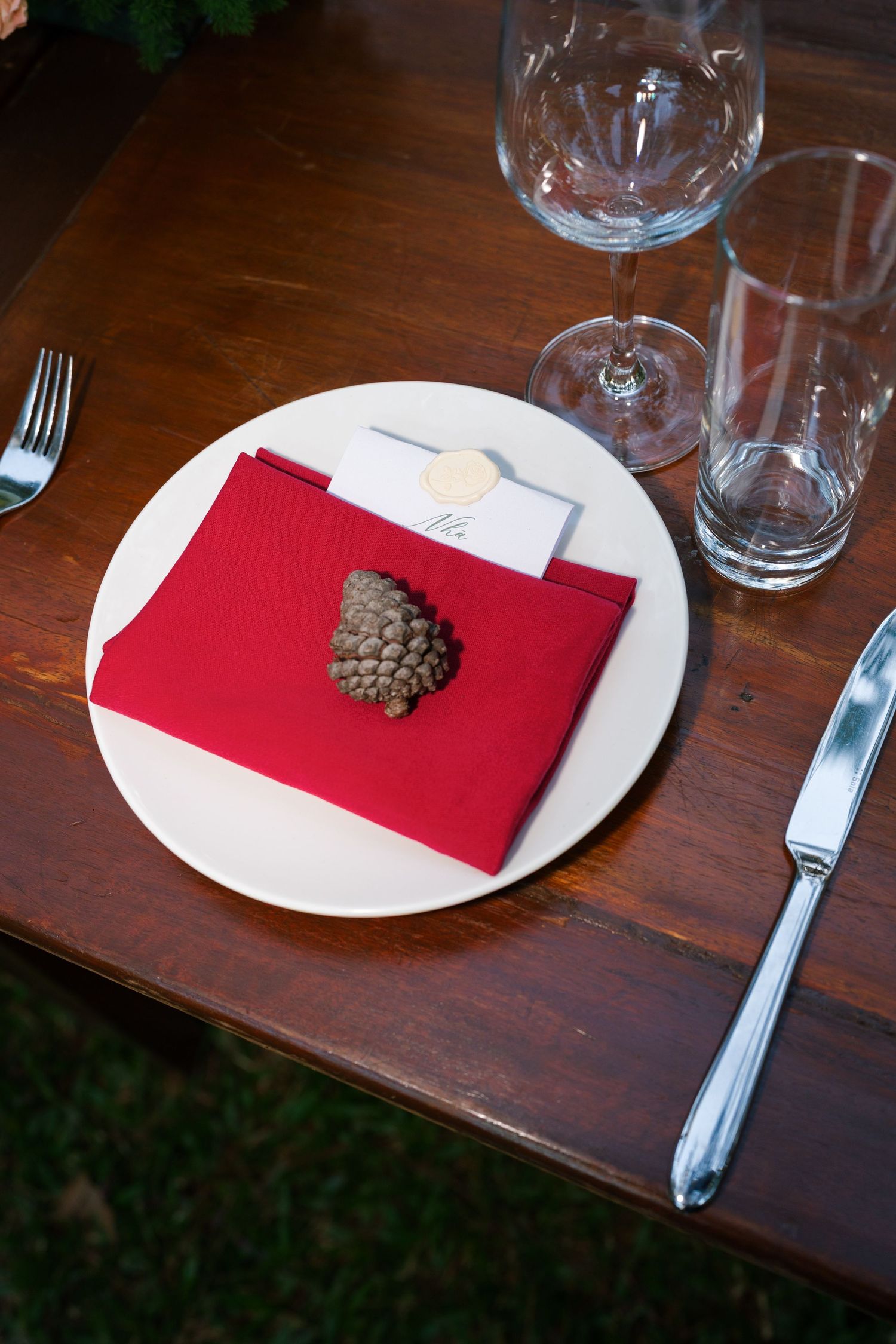 Elegant wedding table setting with red napkin, pine cone accent, place card, and crystal glassware.