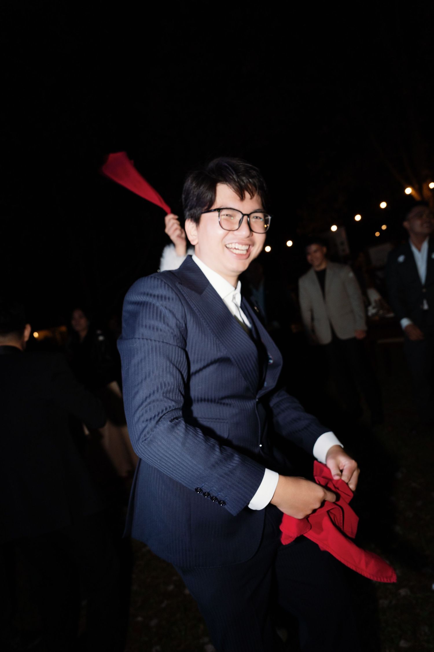 Smiling man in navy suit holding red fabric at lively outdoor evening wedding celebration.