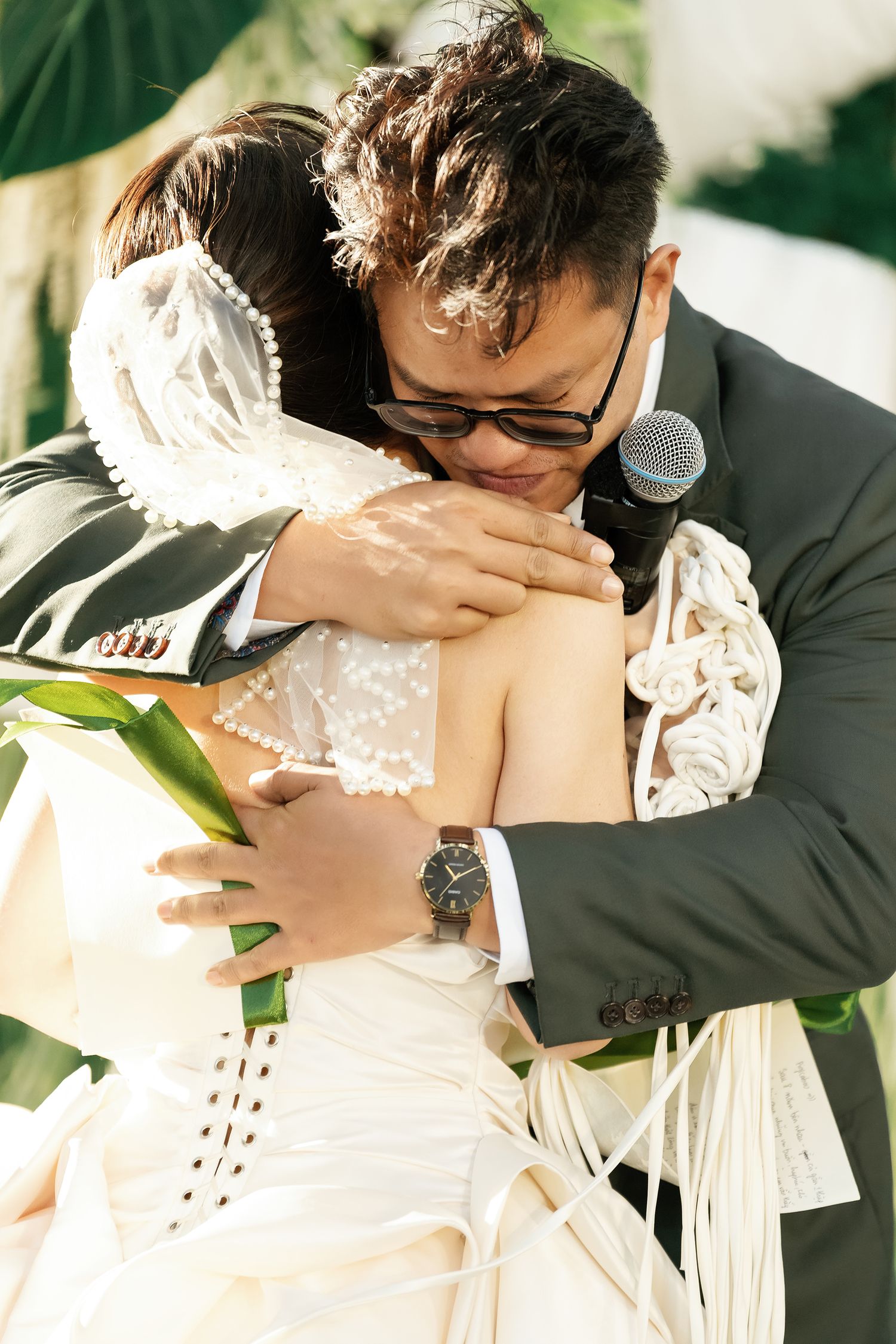 Emotional groom embraces bride during outdoor wedding ceremony while holding microphone.