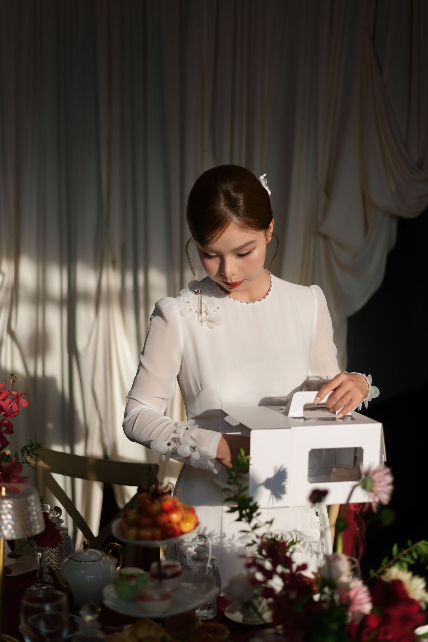 Young woman in white dress and gloves opening a gift box at an elegantly decorated table with flowers.