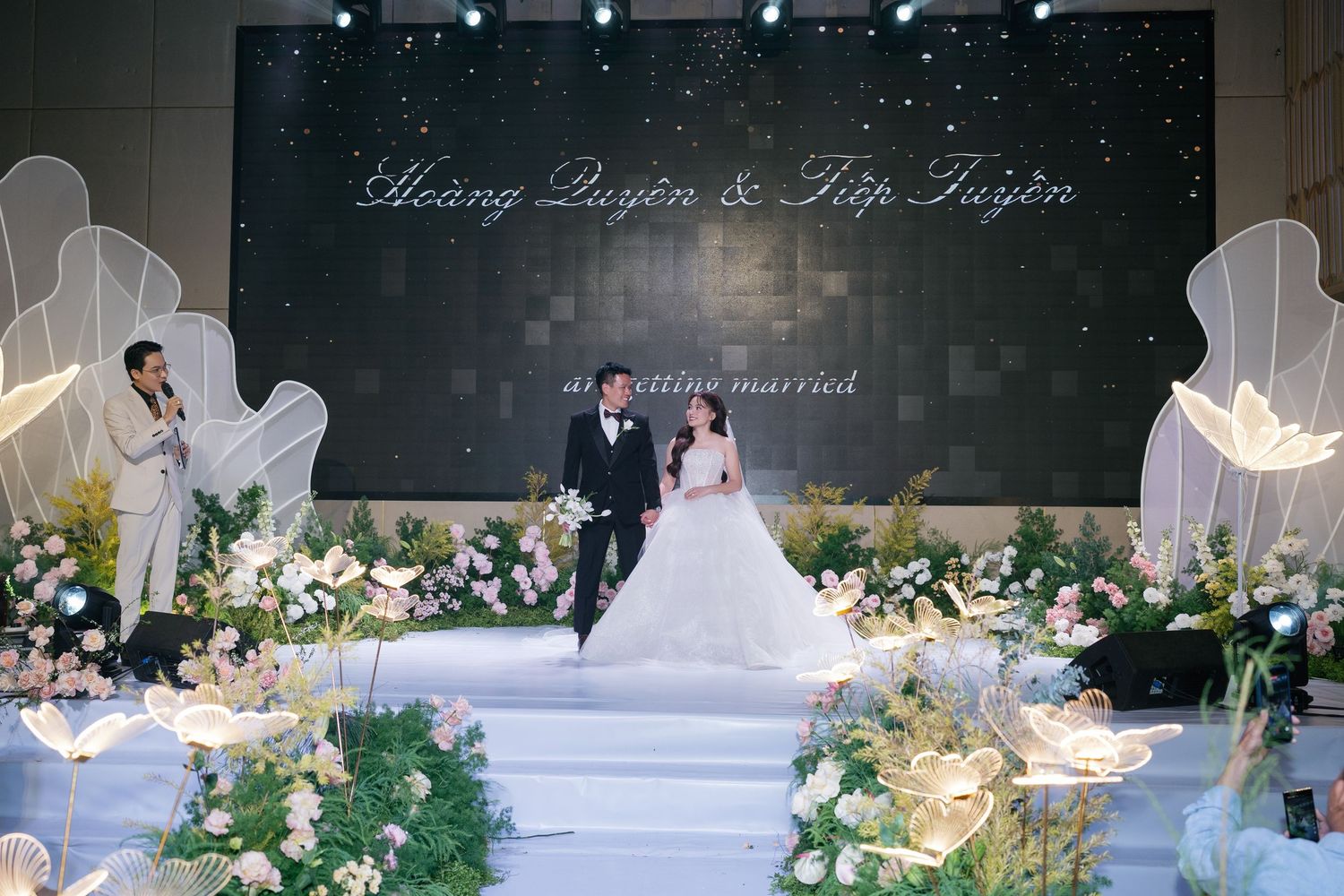 Elegant Vietnamese wedding ceremony with bride and groom on floral decorated stage, backdrop reading Hoang Quyen & Hip Tuyen.