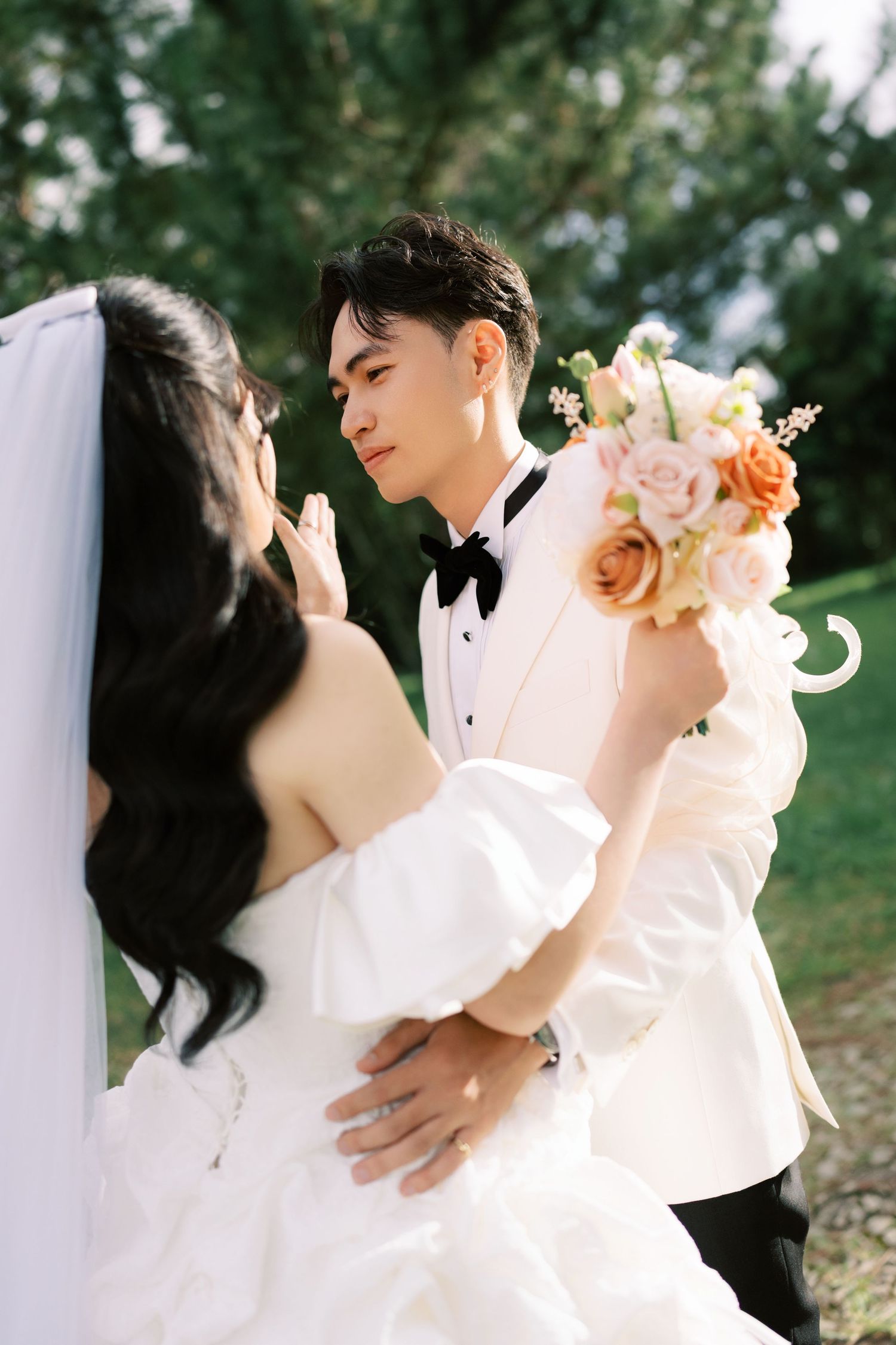 A groom in a white tuxedo holds a floral bouquet while embracing his bride in a white gown outdoors.